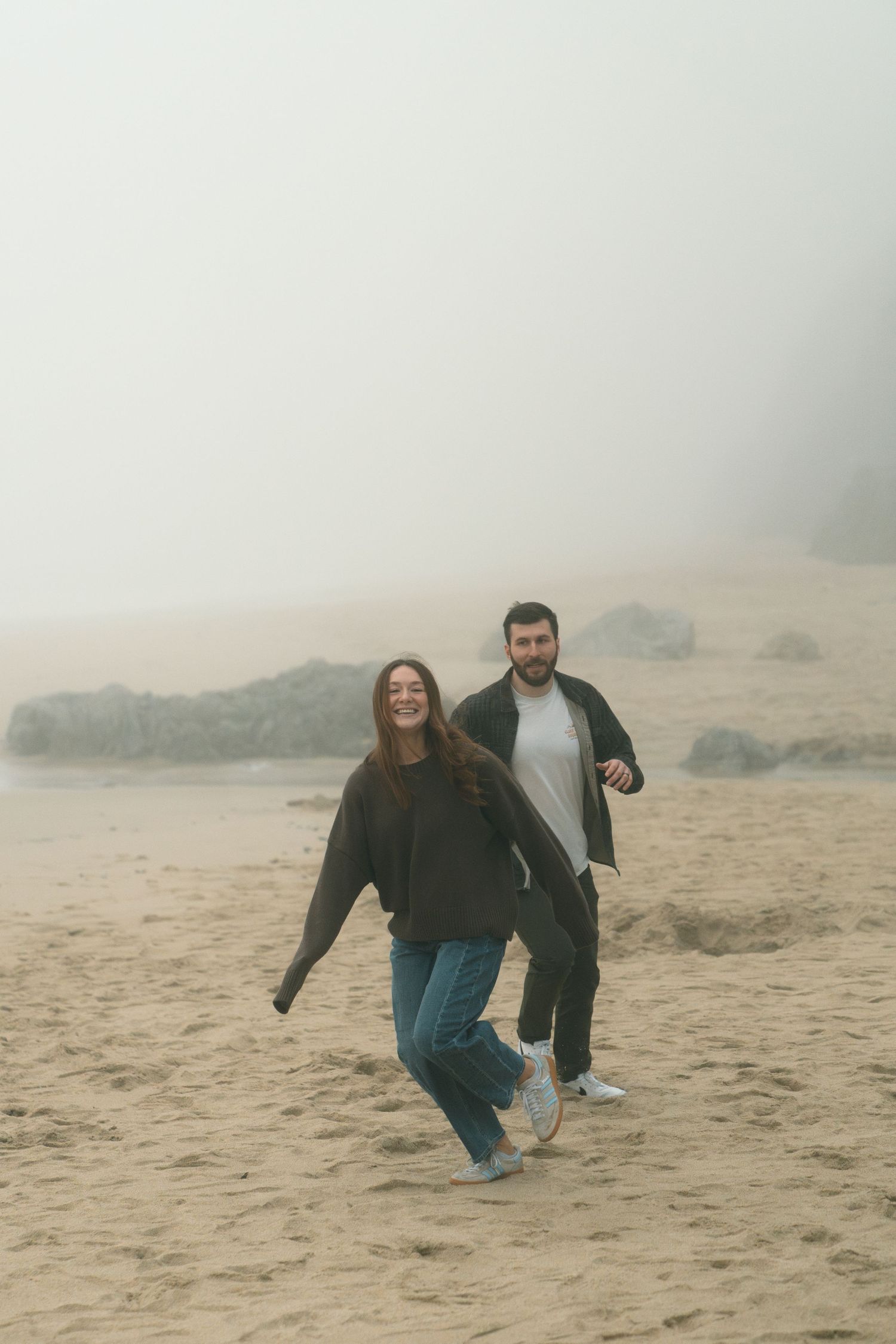 Two people playfully running and laughing on a foggy beach with rocks in the background.