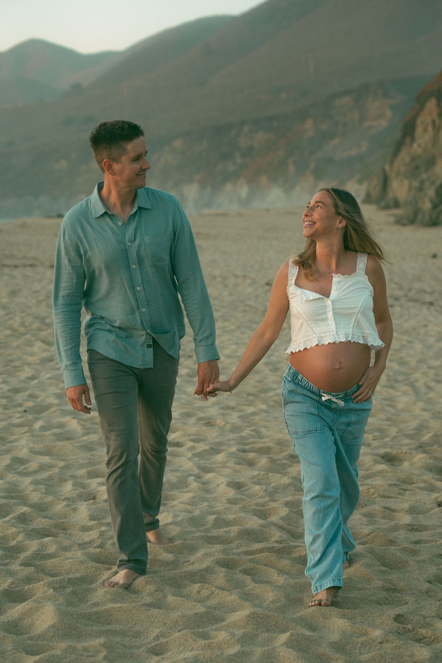 Couple walking hand in hand along a scenic beach with cliffs in the background during golden hour.