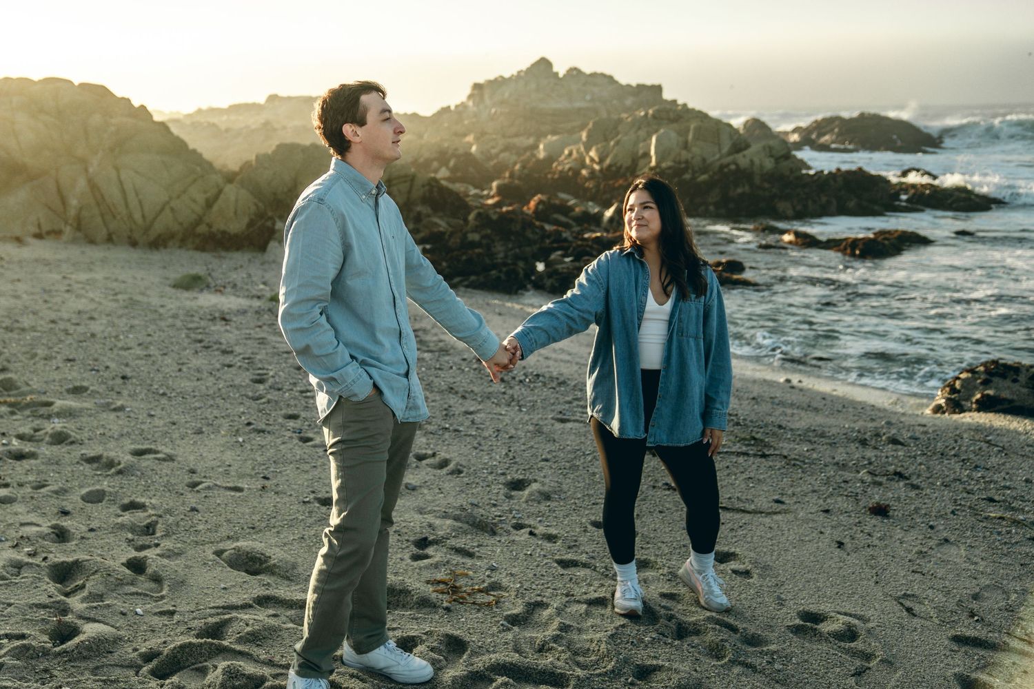 A couple holding hands and walking along a scenic rocky beach at sunset.