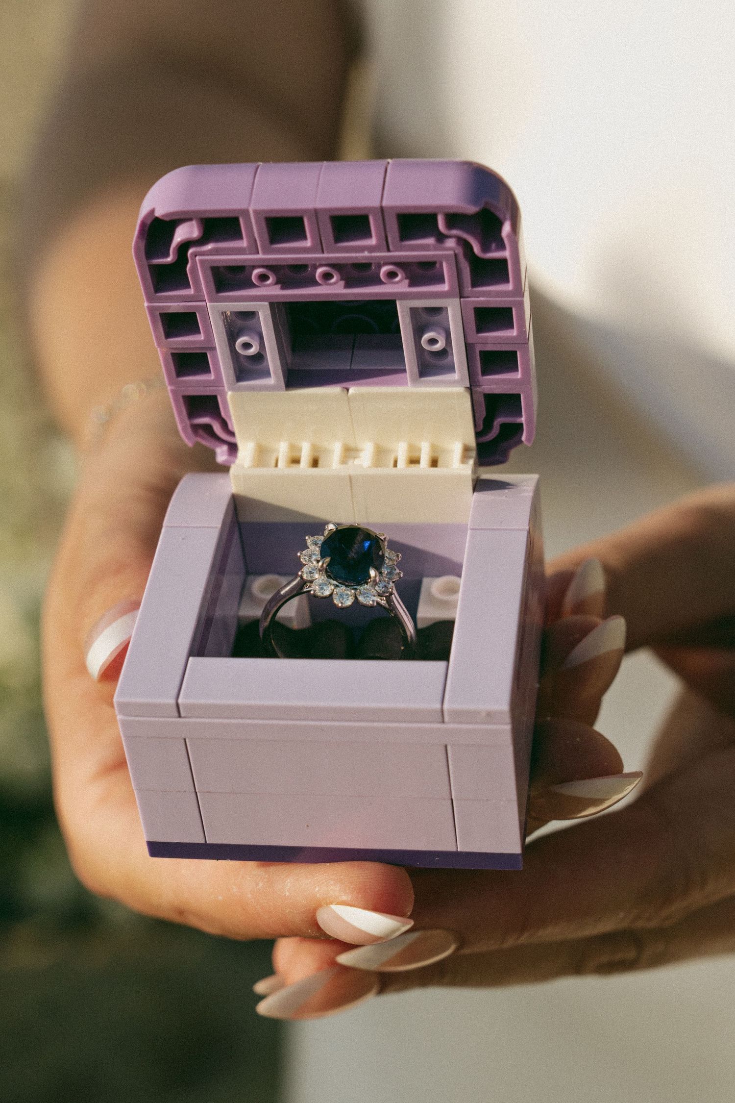 Hand holding a purple LEGO ring box containing a stunning sapphire and diamond halo engagement ring.