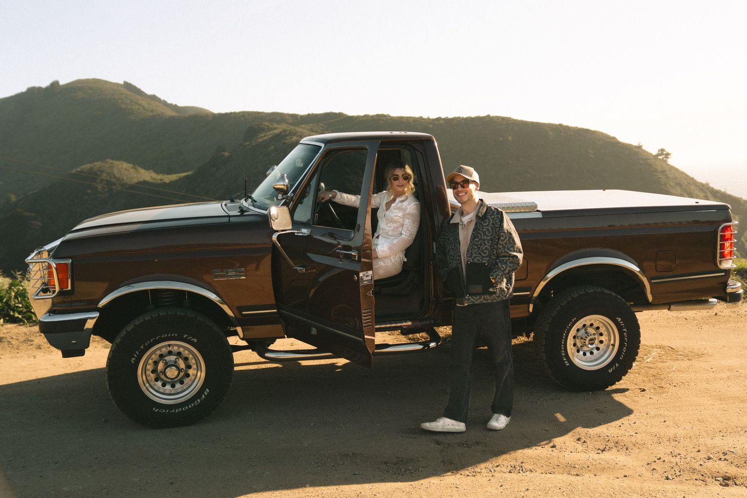 Two people posing with a classic brown Ford F-150 pickup truck on a scenic mountain dirt road.