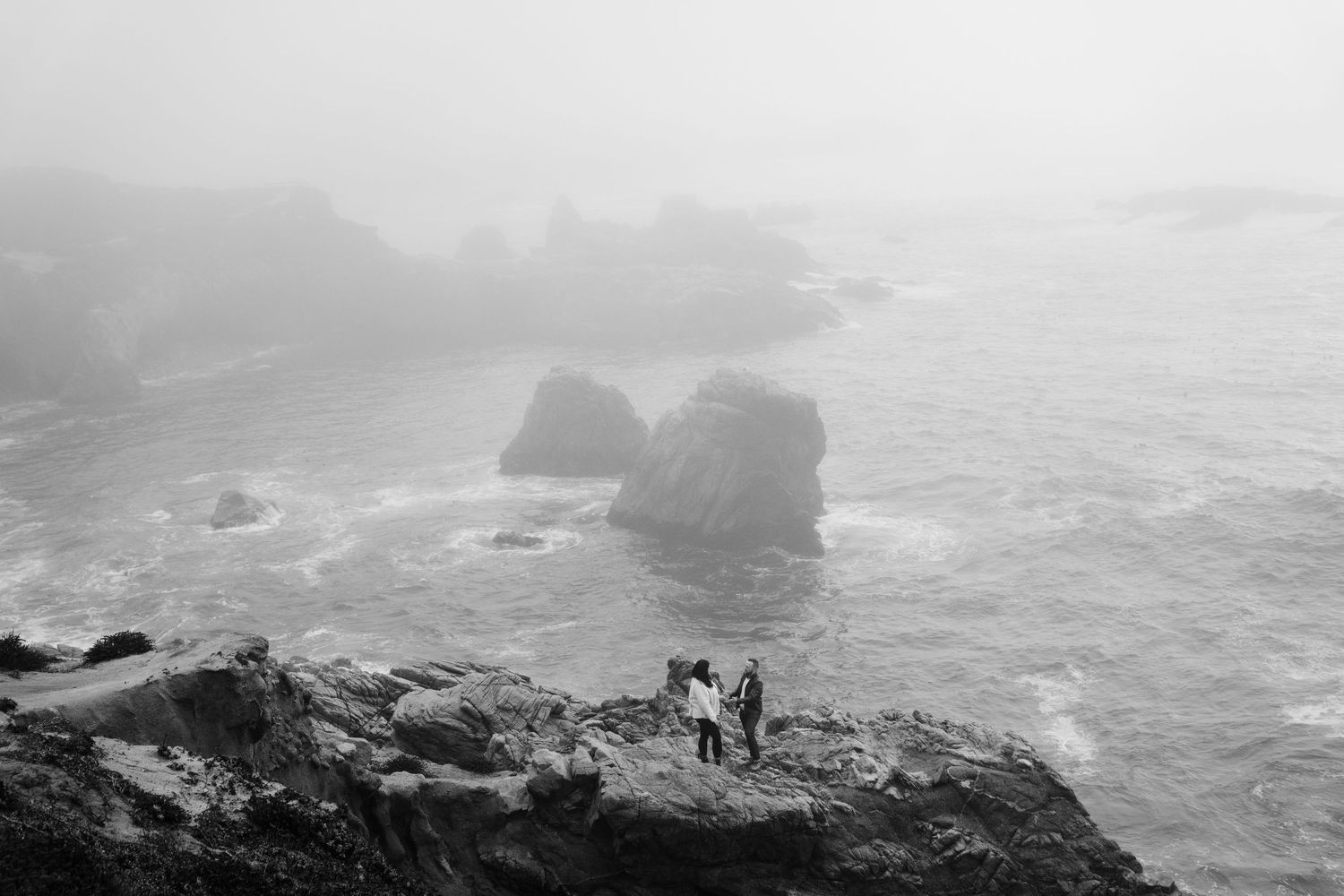 Dramatic black and white aerial view of rugged coastline with waves crashing against rocky cliffs in misty conditions.