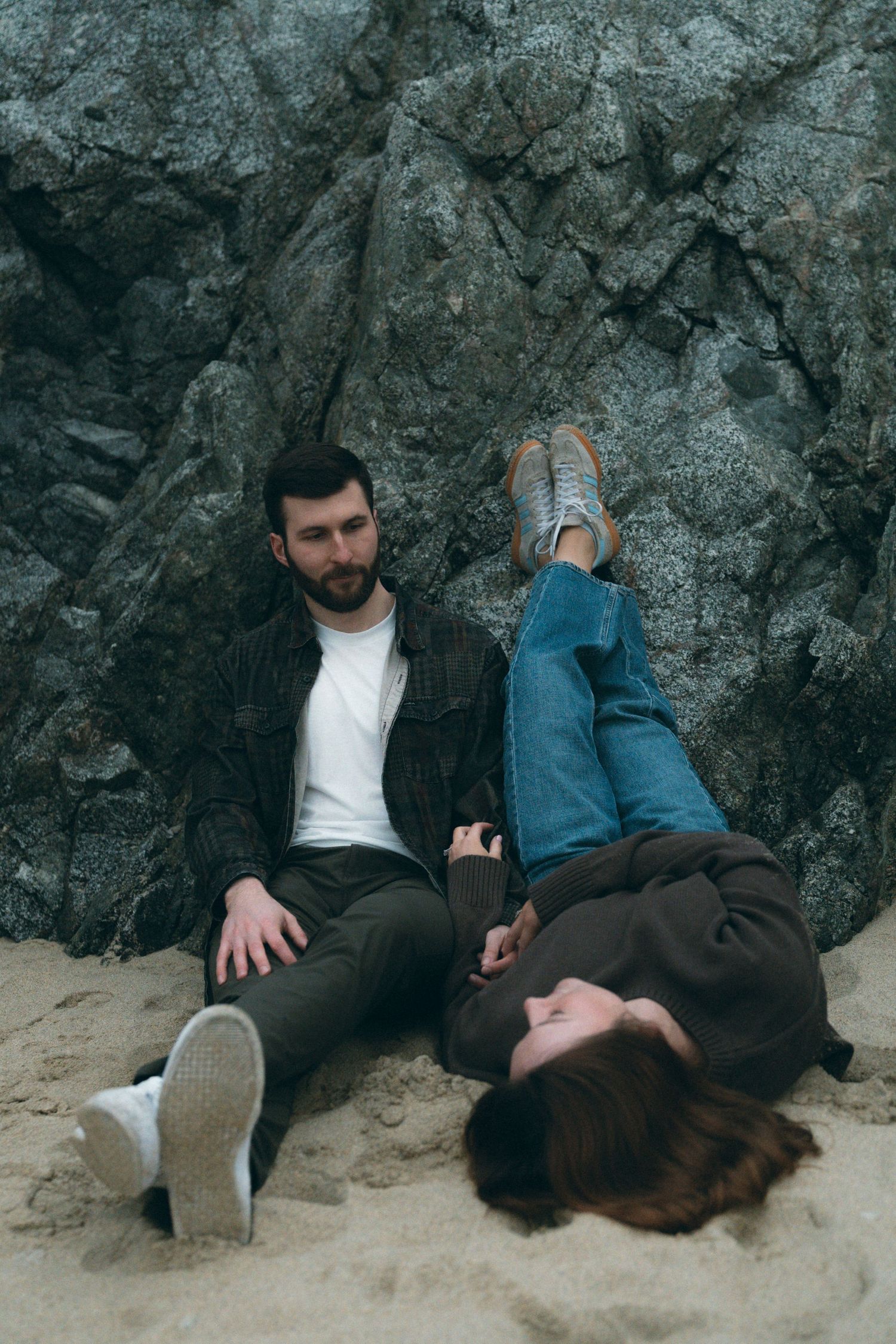 A couple relaxing against dark rocky cliffs on a sandy beach.