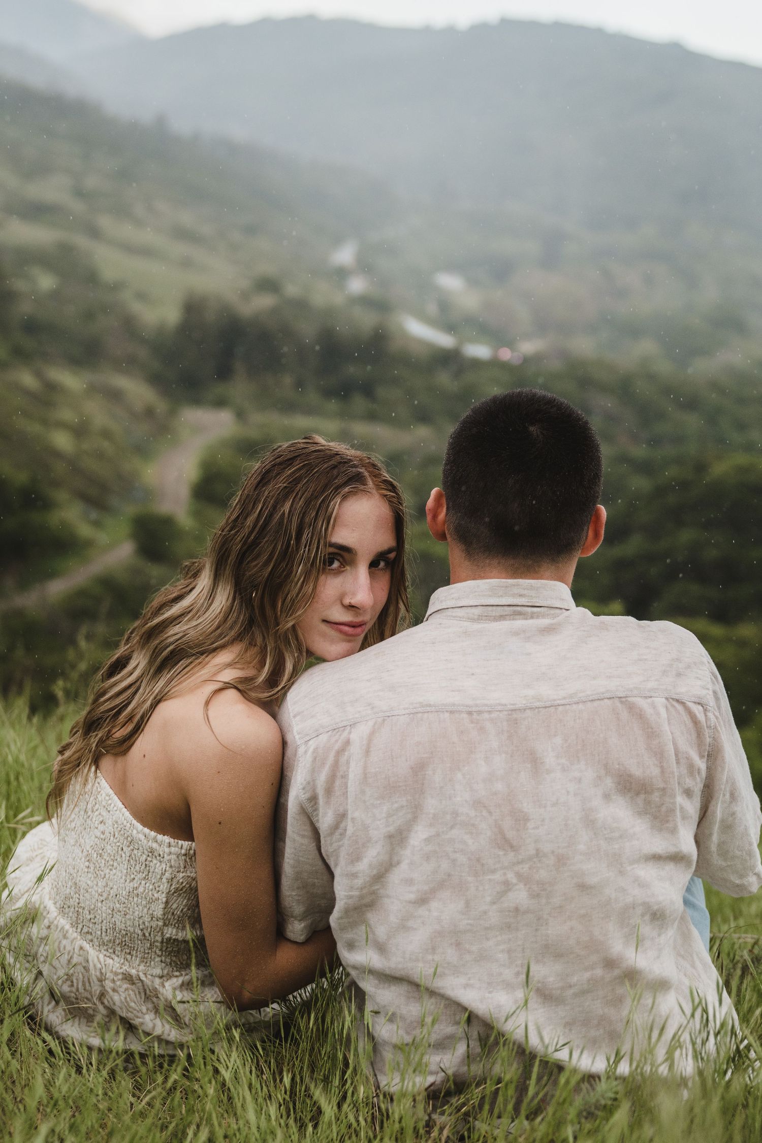 A couple sits together in tall grass overlooking a mountain valley.