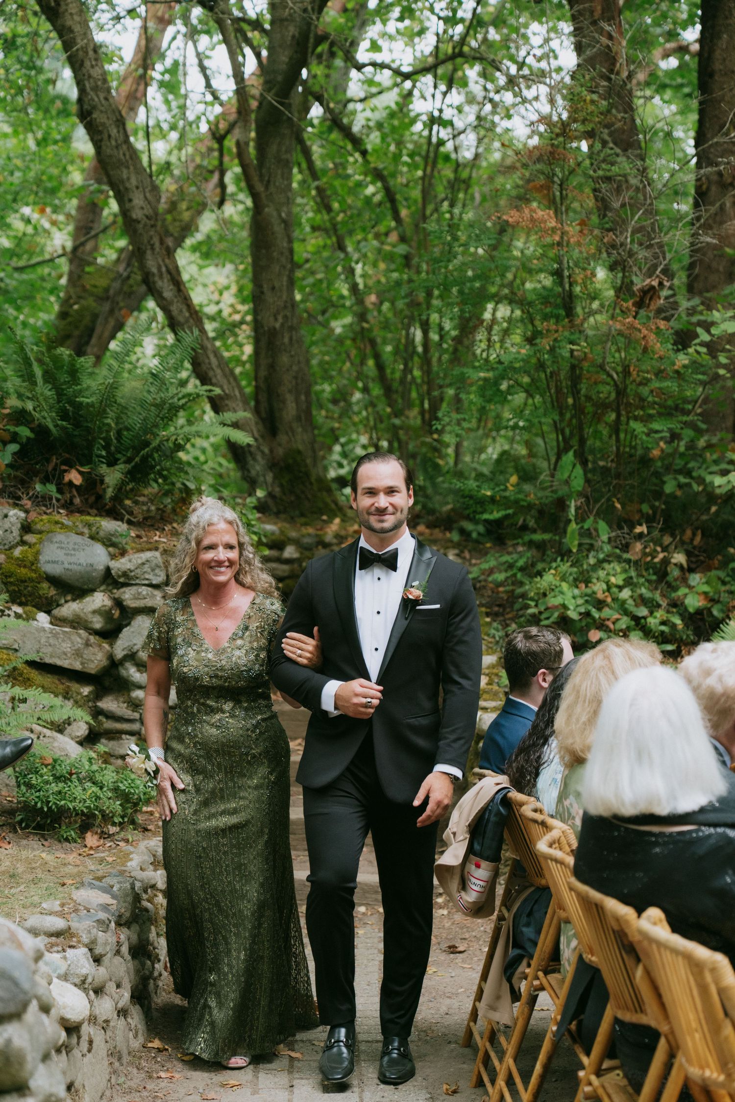 Wedding ceremony guests seated on wooden chairs in wooded outdoor setting with stone path.