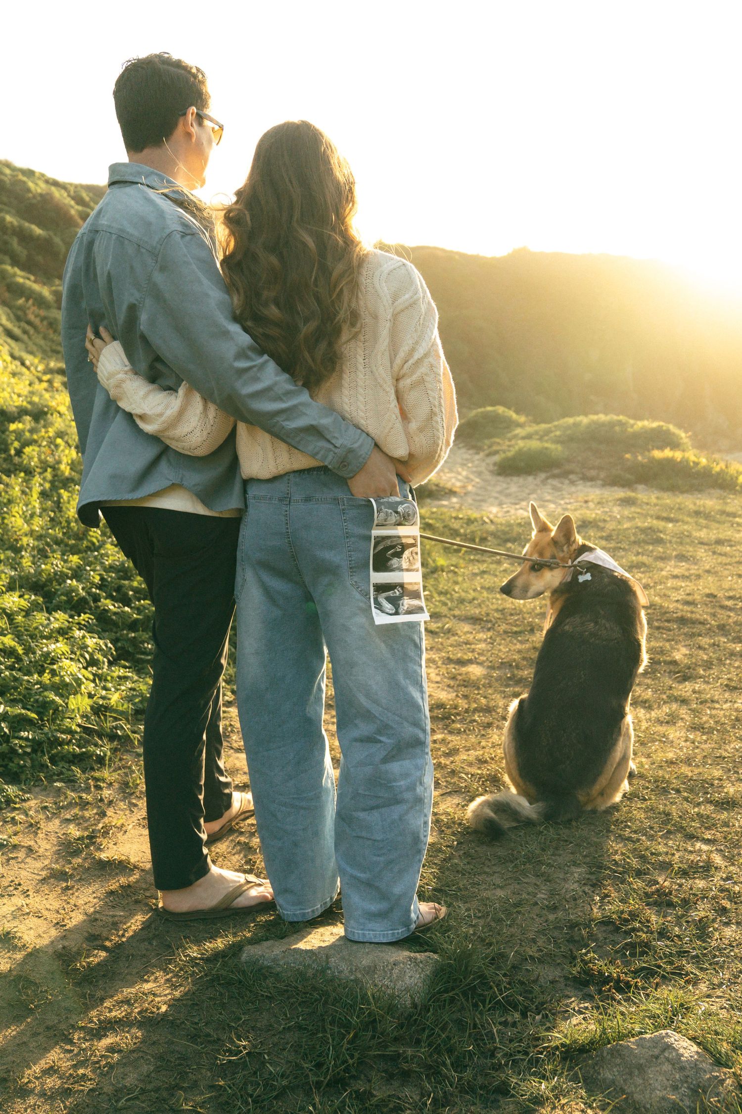 Couple embracing at sunset on a hillside with their dog on a leash, viewed from behind in casual attire.