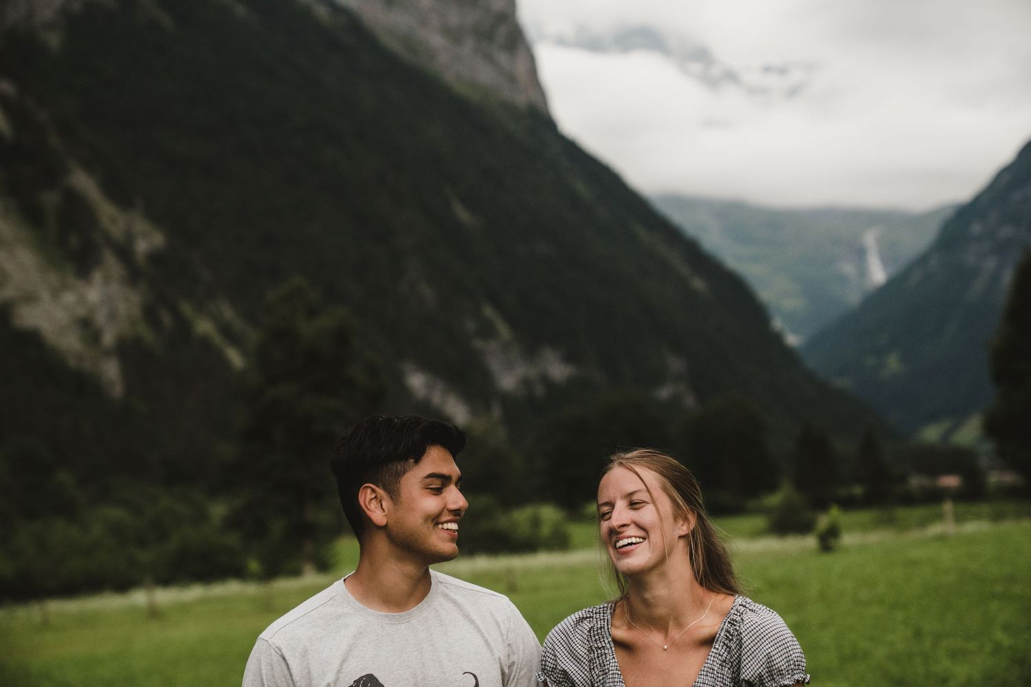 A joyful couple laughing together while standing in a scenic alpine meadow with towering mountains in the background.