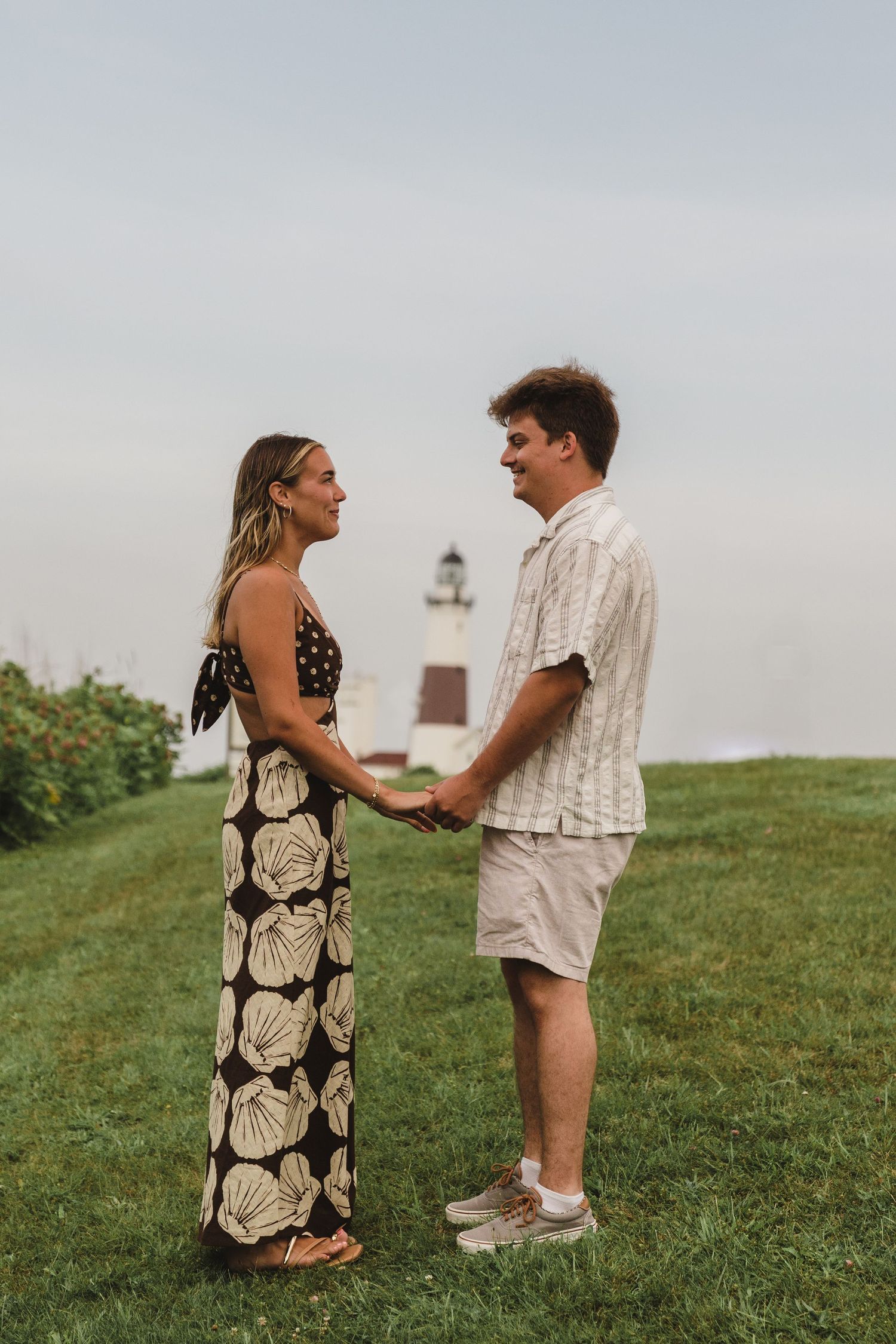 A romantic moment between a couple standing on a grassy hill with a lighthouse visible in the background.