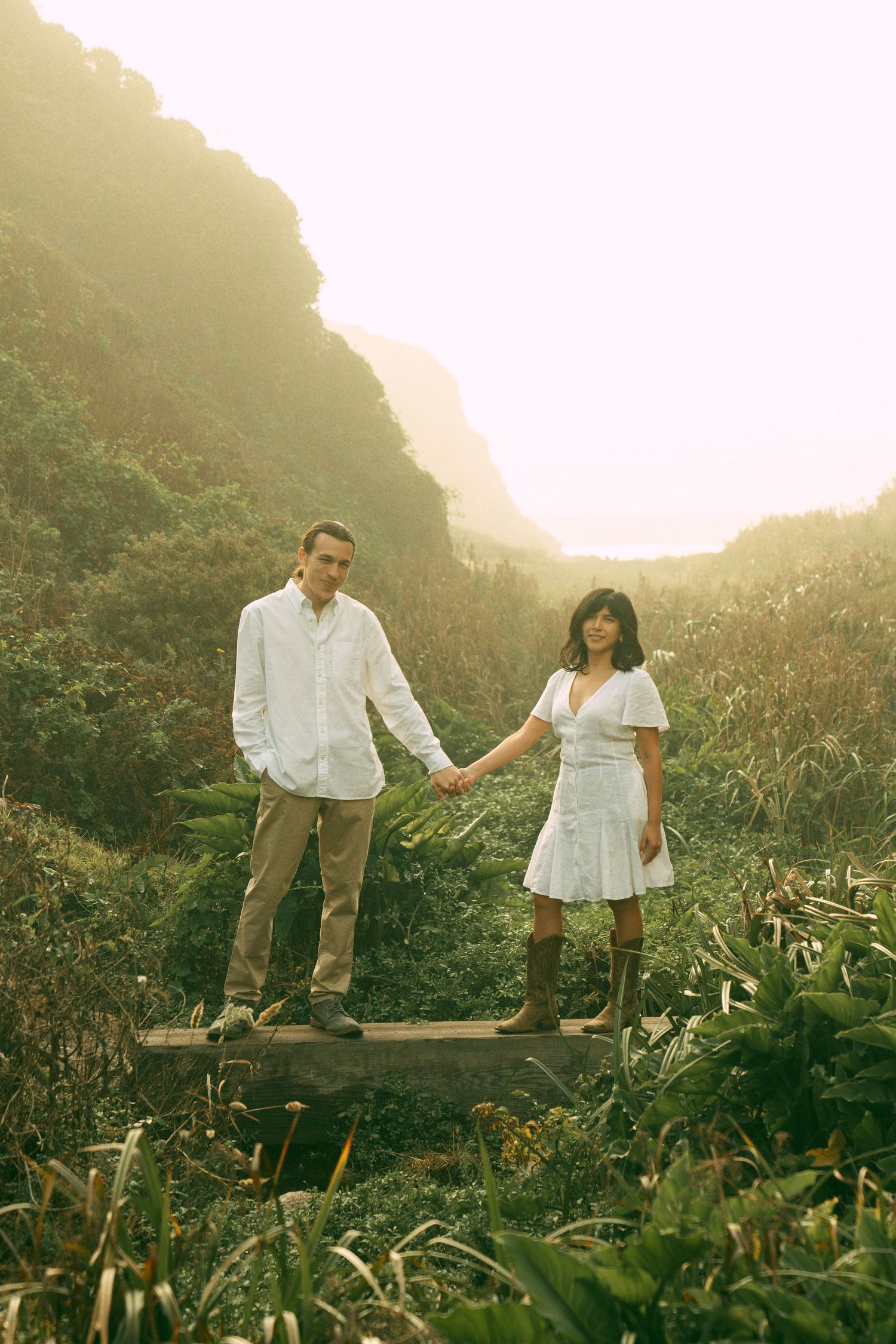 A couple in white outfits walks together through a misty mountainous landscape at golden hour.