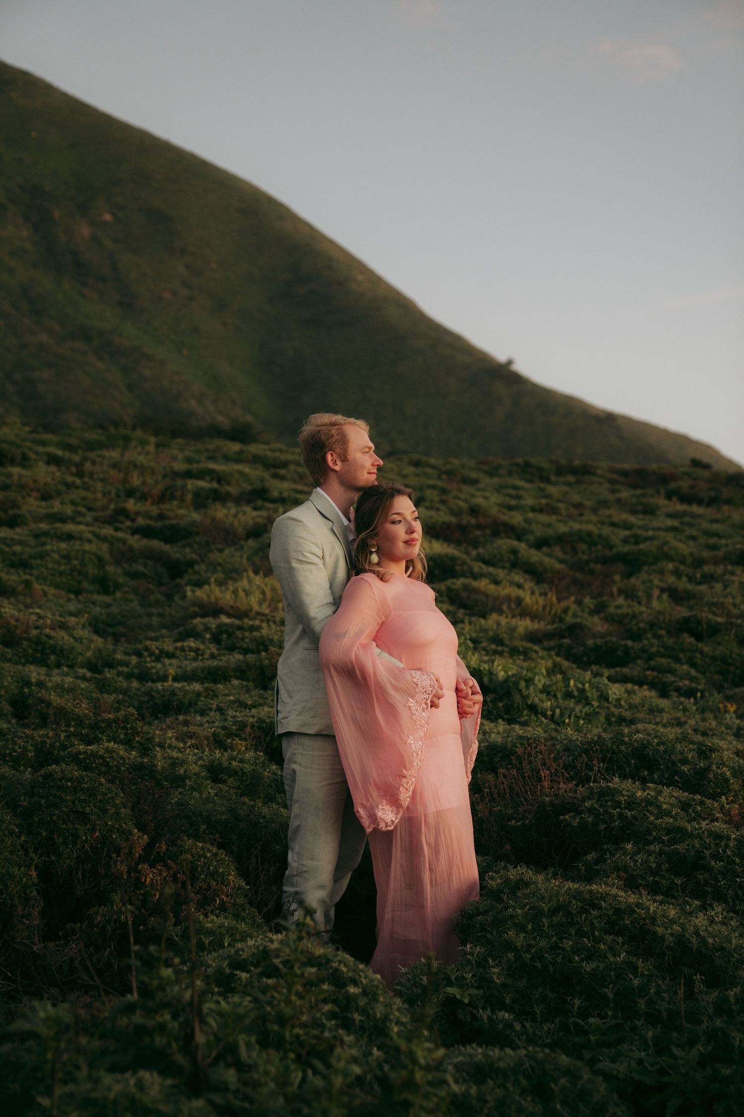 Couple embracing on a lush green hillside at golden hour, woman in pink gown cradling baby bump.