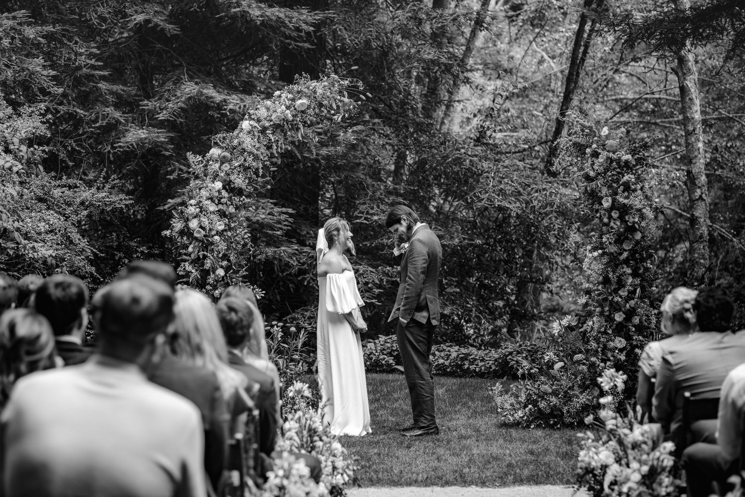 A black and white outdoor wedding ceremony takes place on a wooded path surrounded by lush greenery and seated guests.