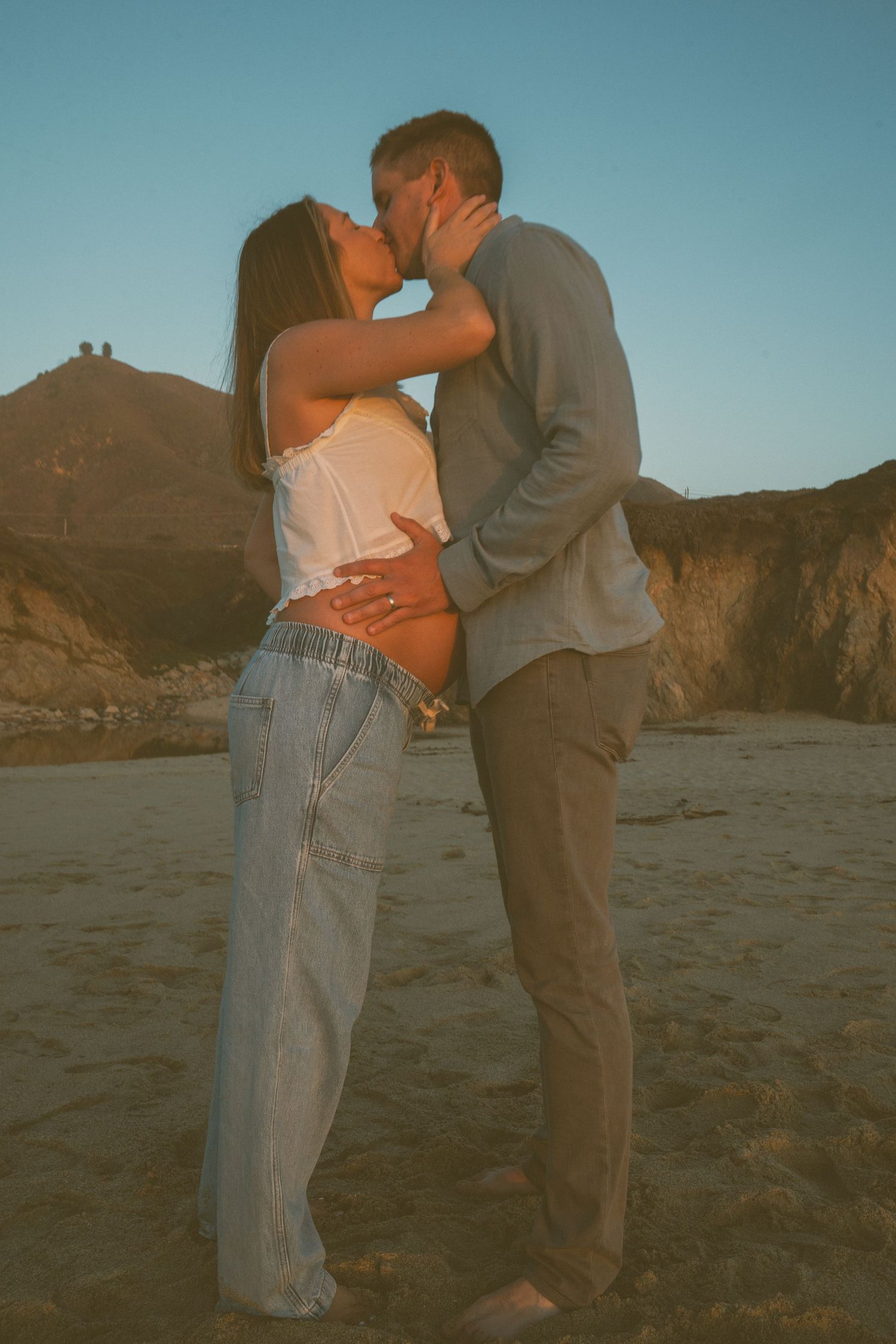 A romantic couple embraces on a beach at sunset with mountains visible in the background.