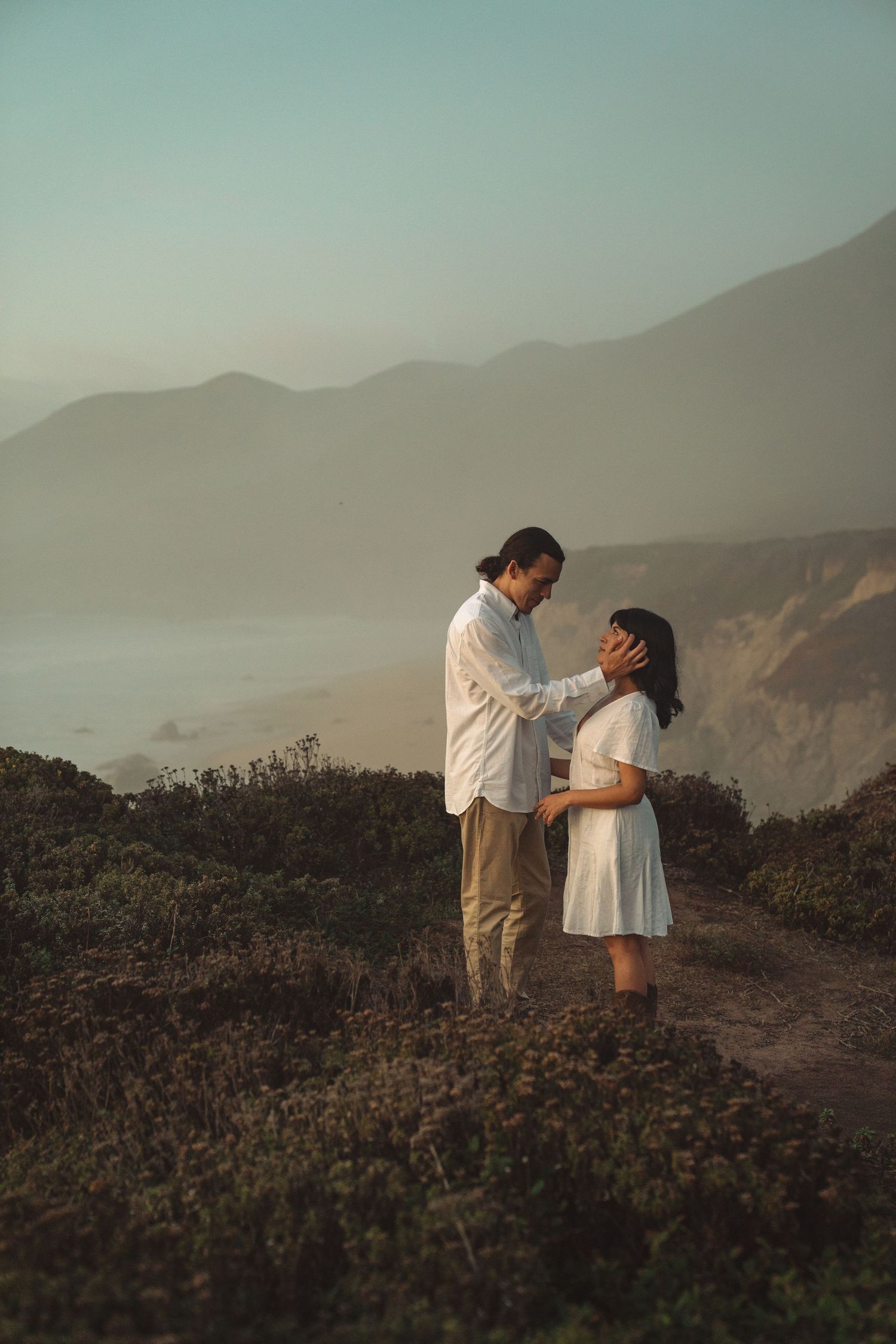 A couple shares a romantic moment on a misty mountain trail at sunset with coastal views in the background.