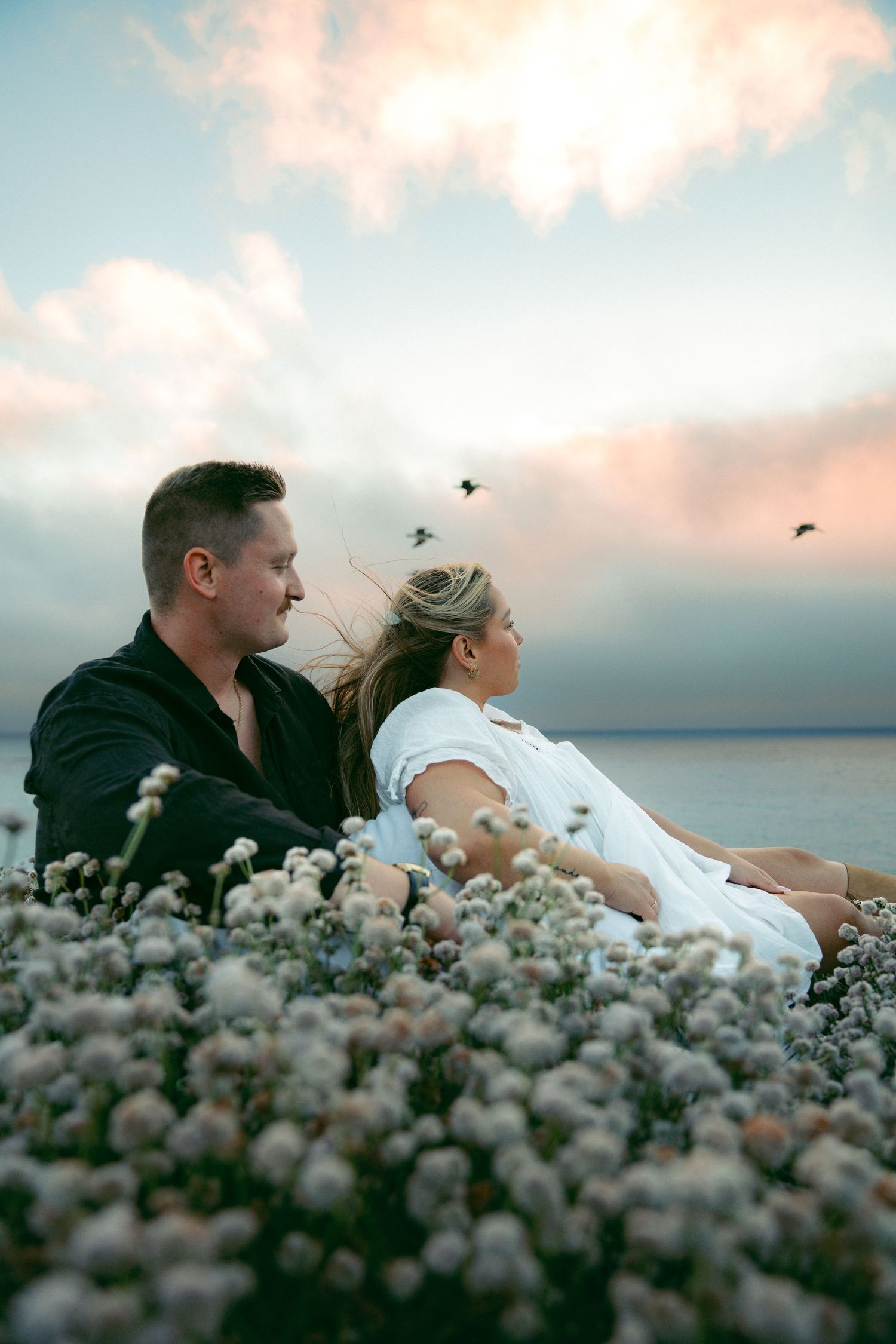 A couple sits among wildflowers at sunset while birds fly overhead against a pastel sky.