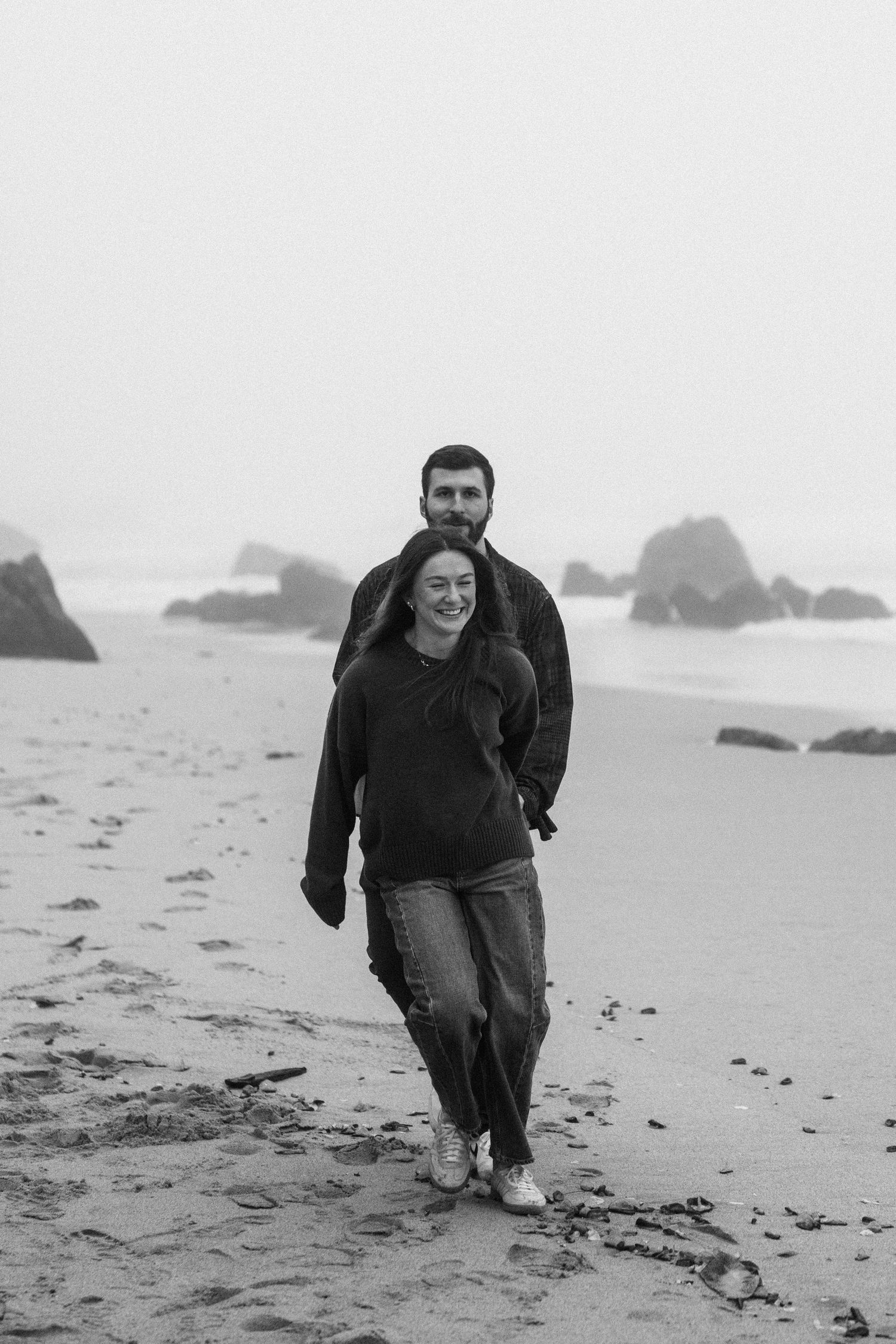 A couple taking a romantic stroll on a rocky beach with sea stacks visible in the misty background.