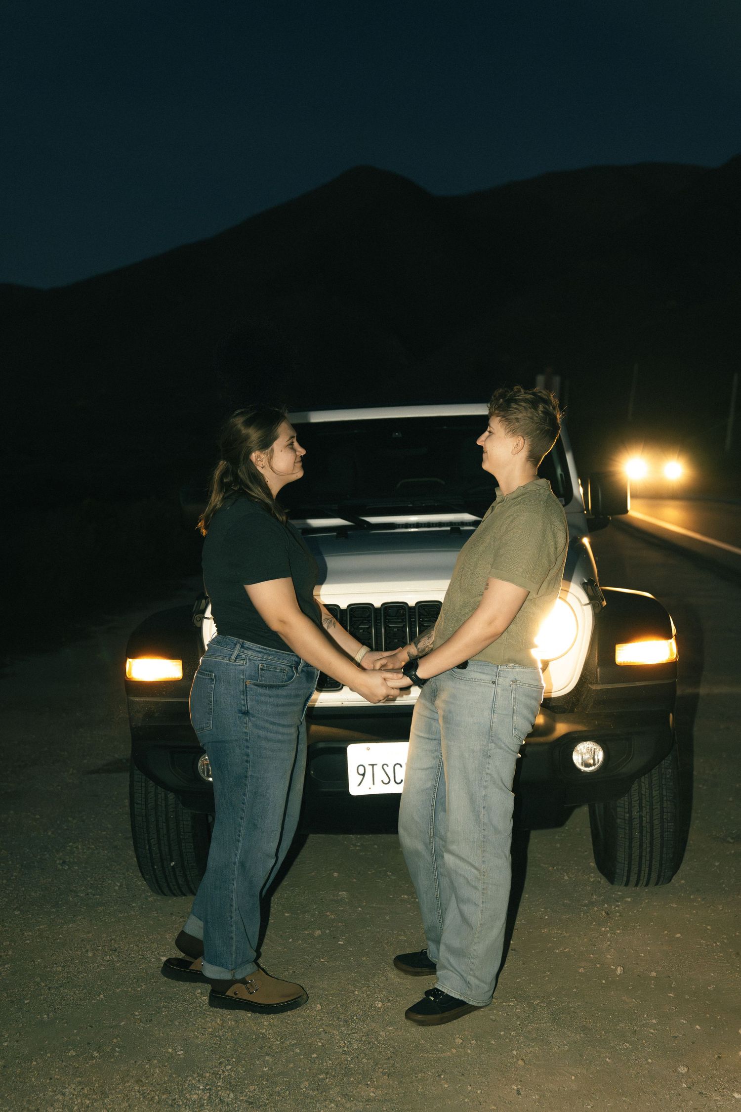 A couple holds hands in front of a car with headlights on at night against a mountain backdrop.