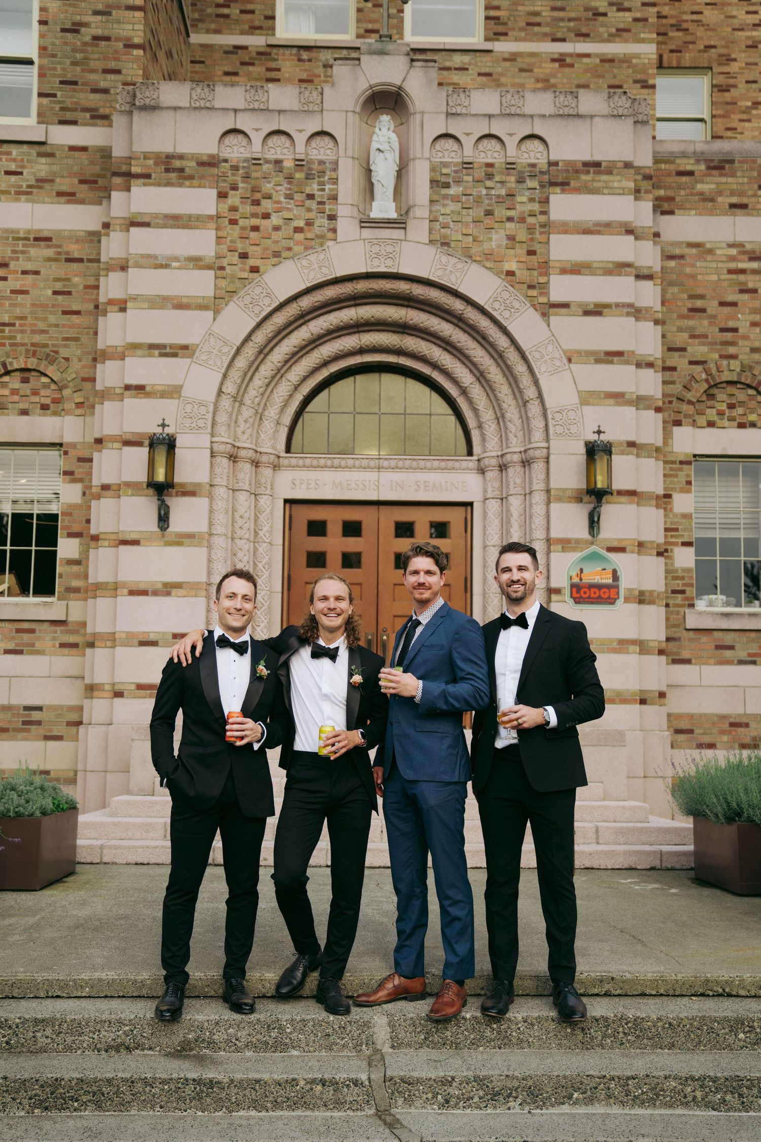 Groomsmen in tuxedos pose with drinks outside historic brick building with arched doorway.