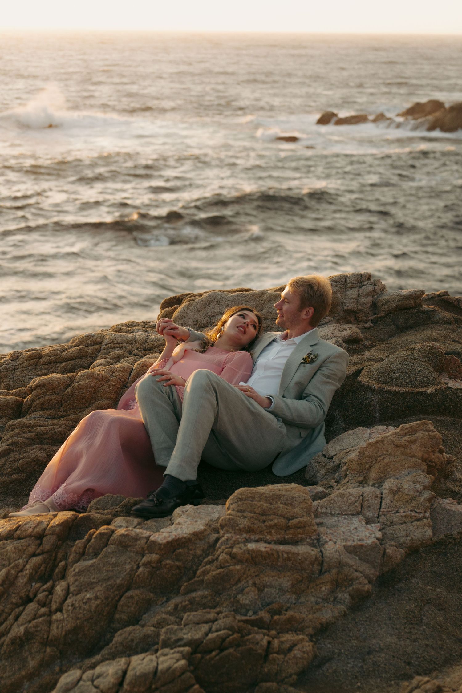 Couple in formal wear relaxing on coastal rocks at sunset with ocean waves in the background.