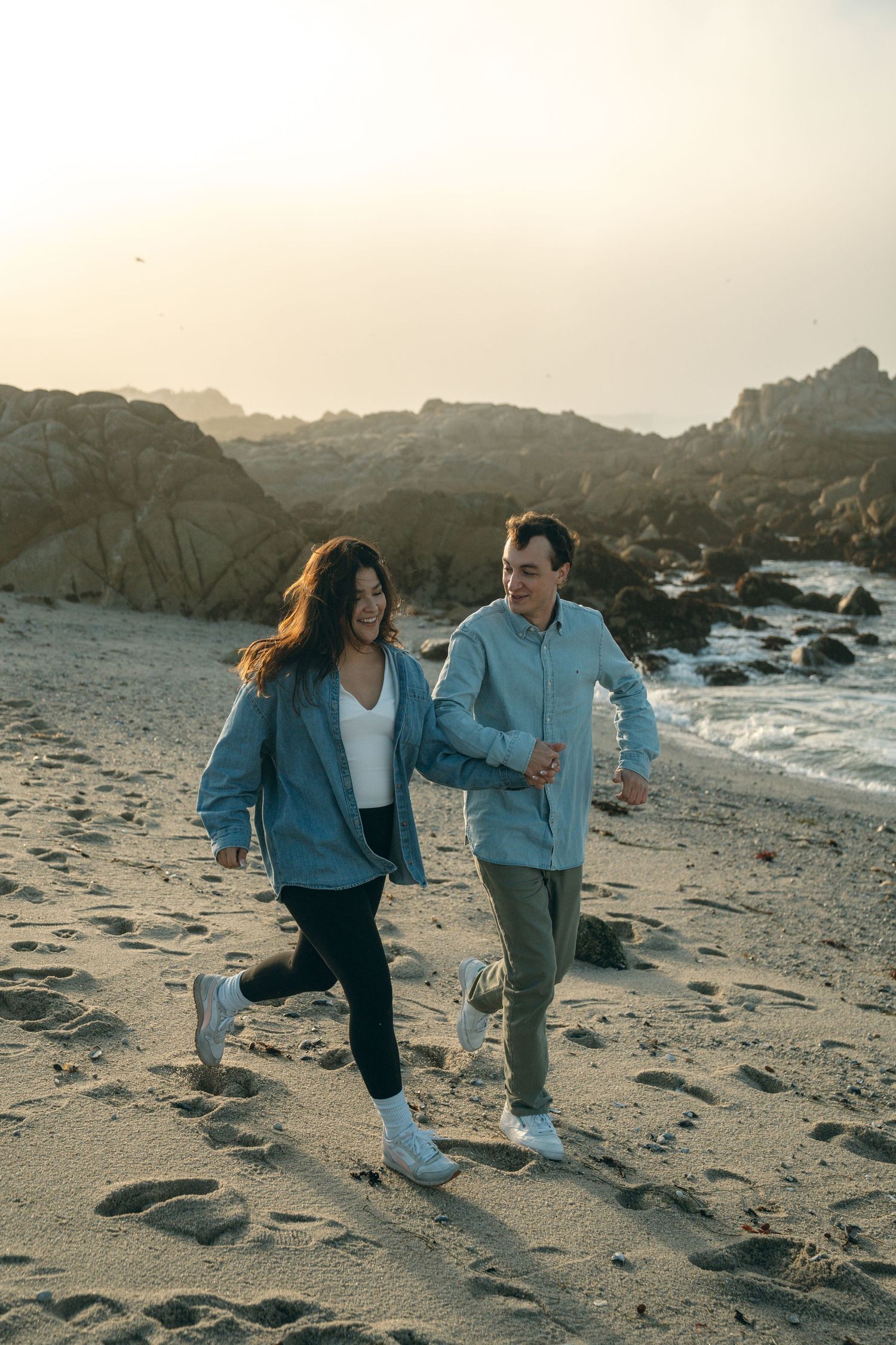 A couple runs playfully along a rocky beach at sunset with matching denim jackets and casual attire.