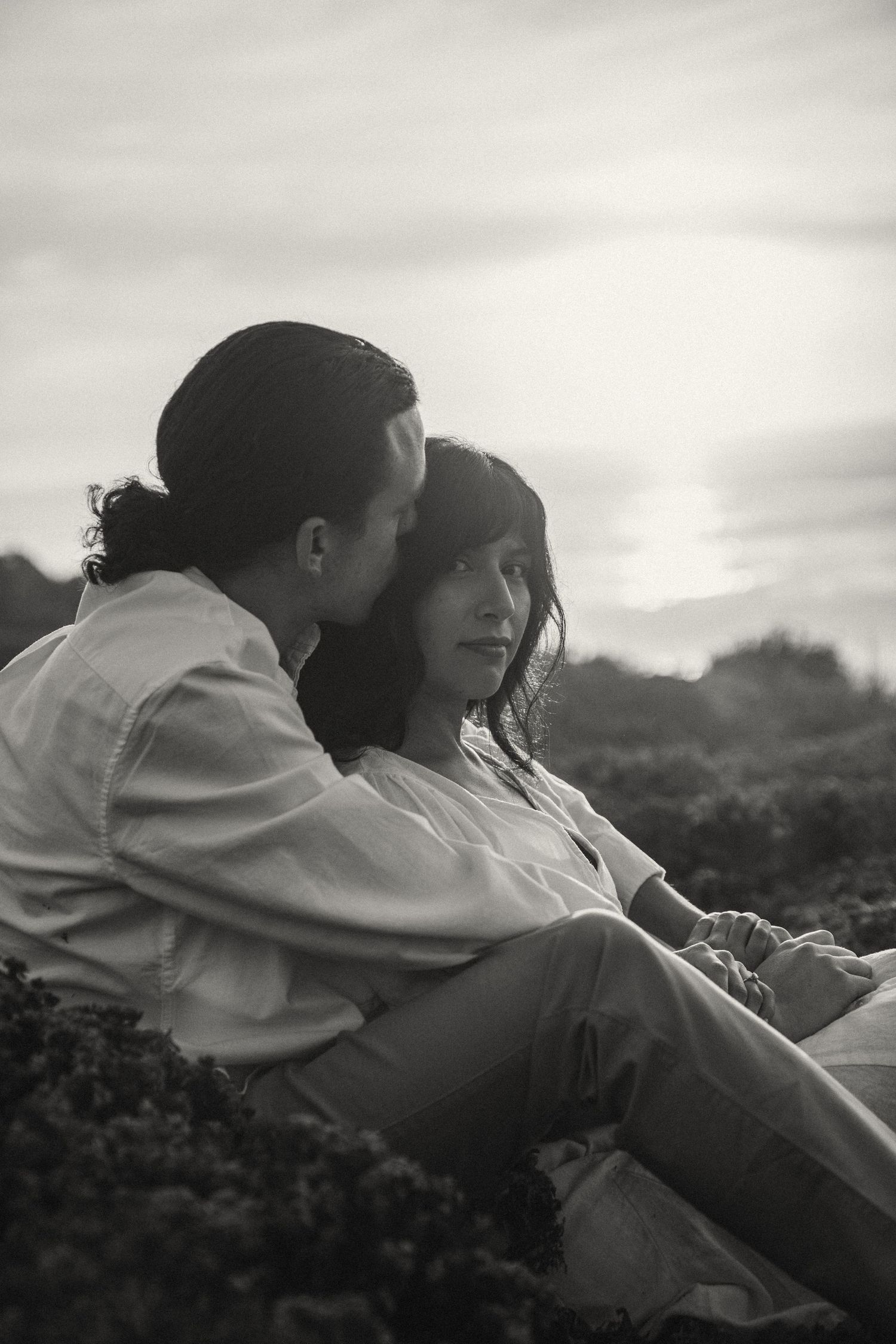 Black and white photograph of a couple sharing an intimate moment on a hilltop.