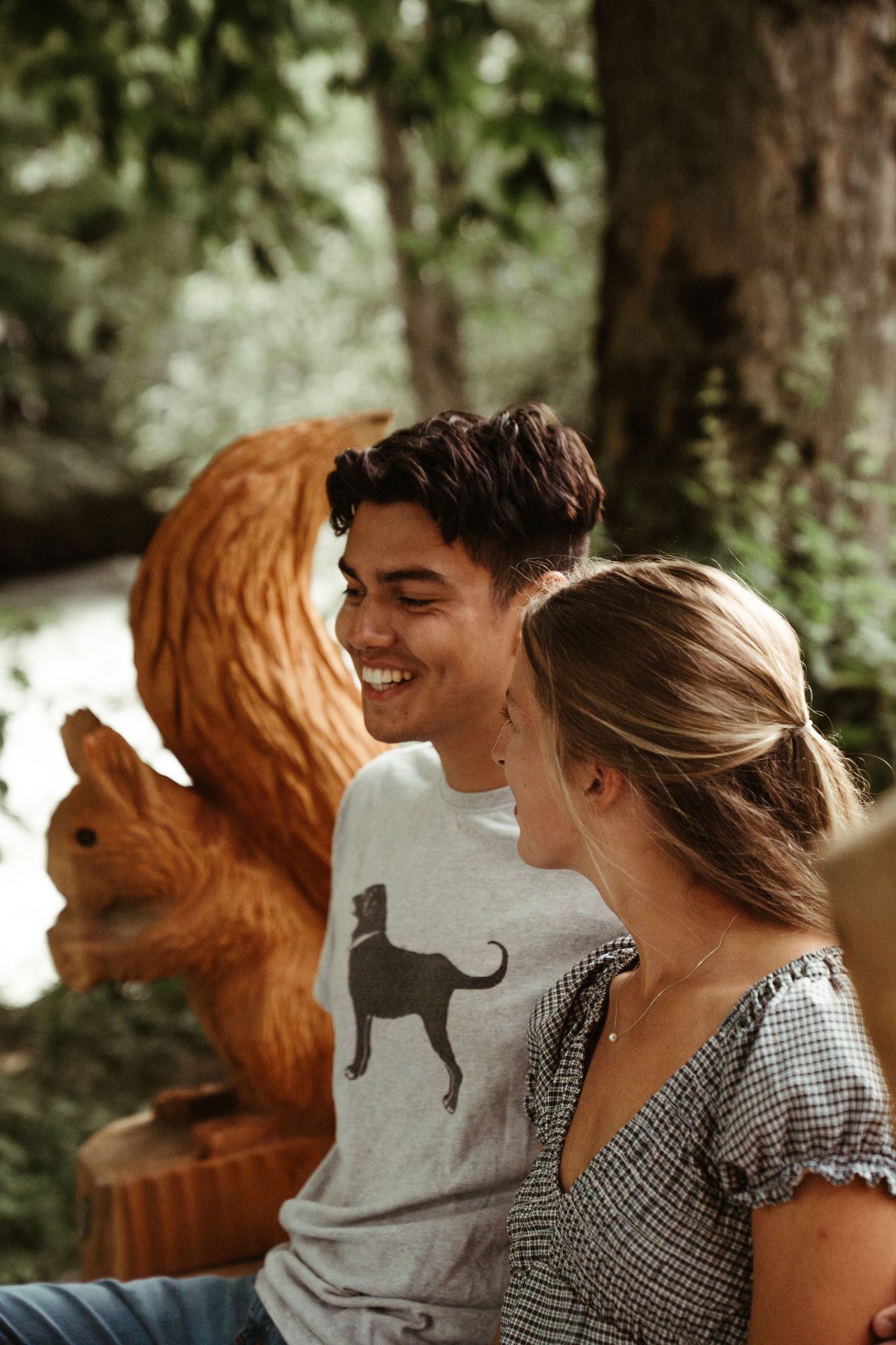 A couple laughs together on a bench near a carved wooden fox sculpture in a garden setting.