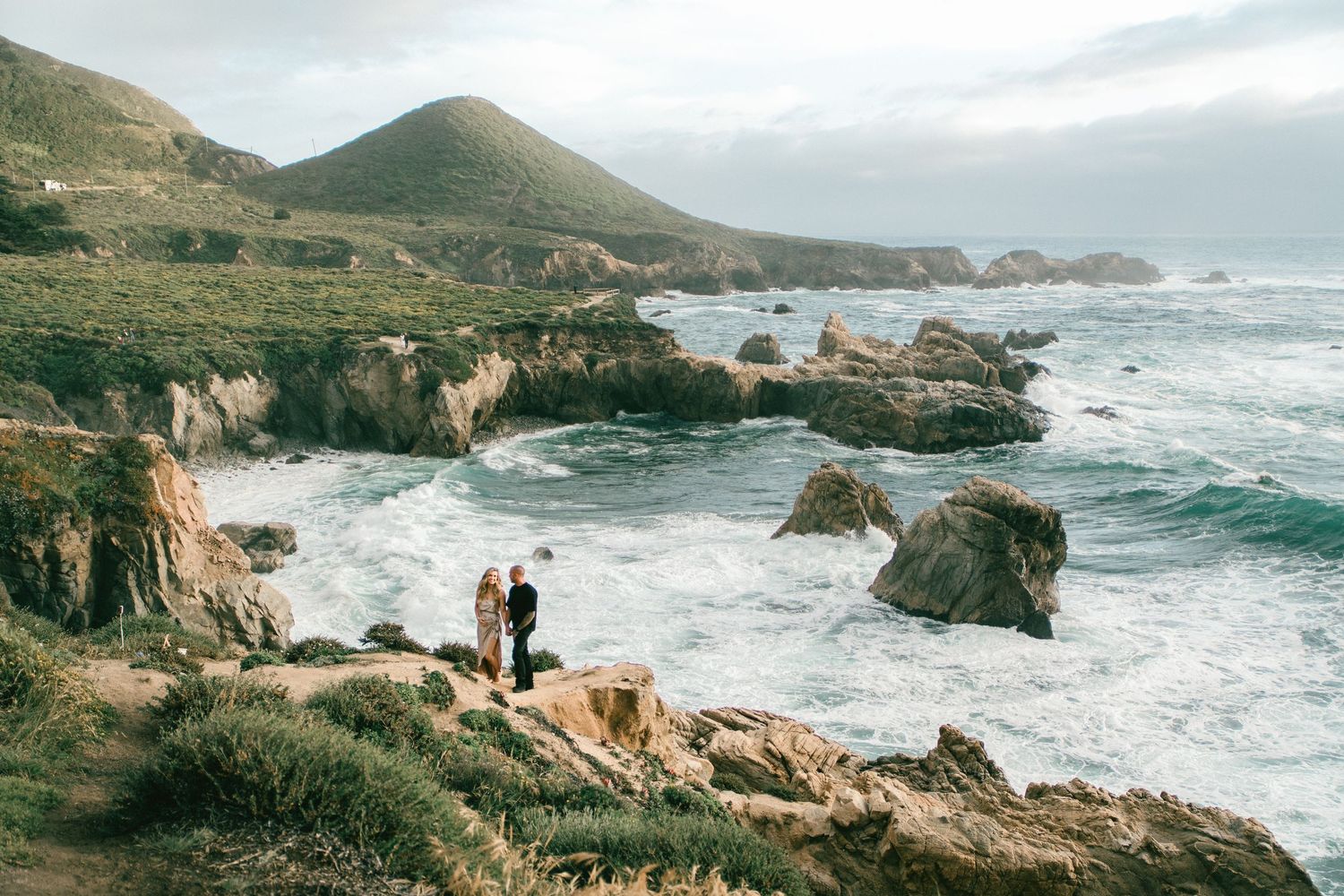 Couple standing on rocky coastal cliffs overlooking dramatic ocean waves with green hills and sea stacks in Big Sur, California.