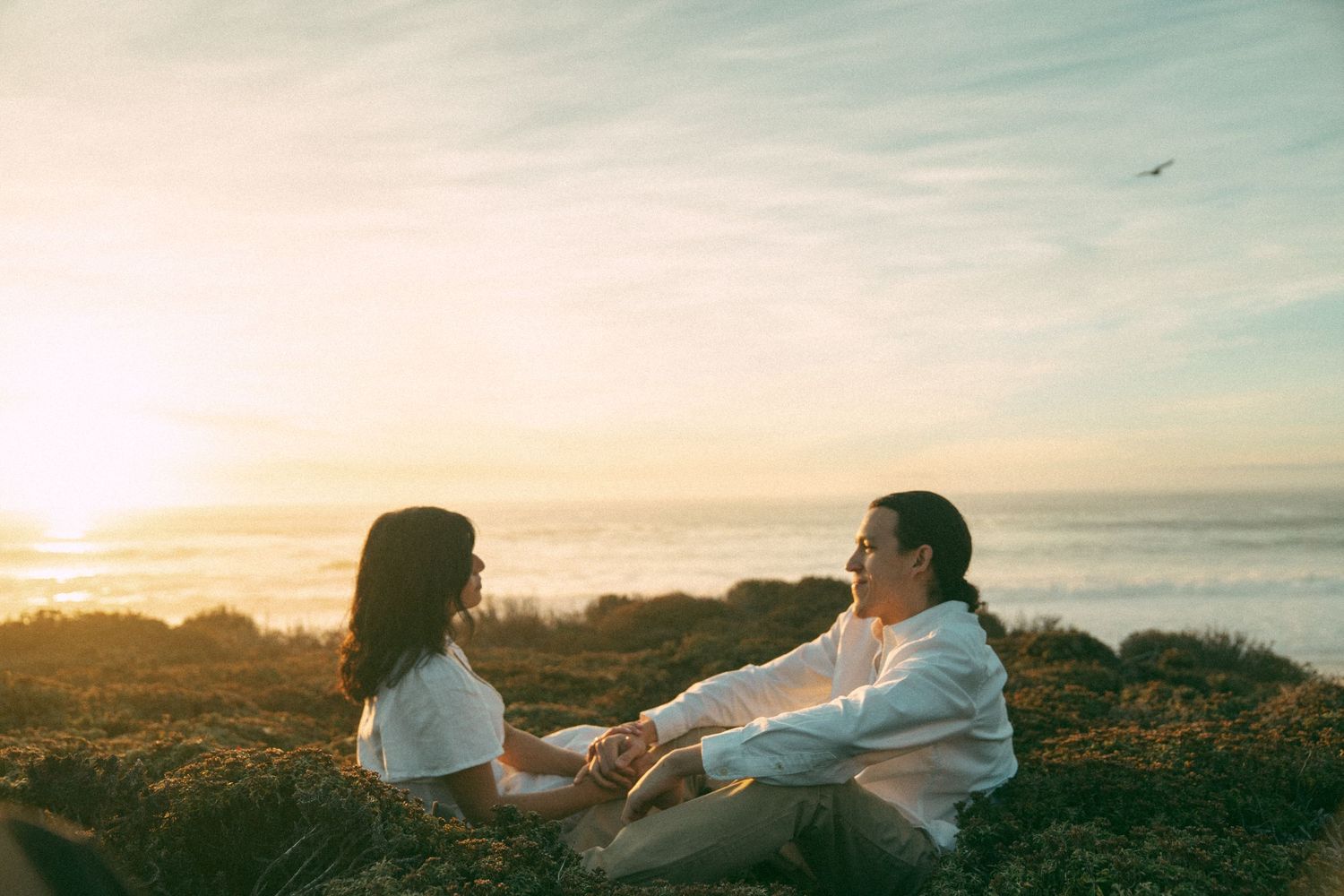 Elopement couple relaxes together in a grassy field during a scenic sunset on Big Sur with clouds streaking across the sky.
