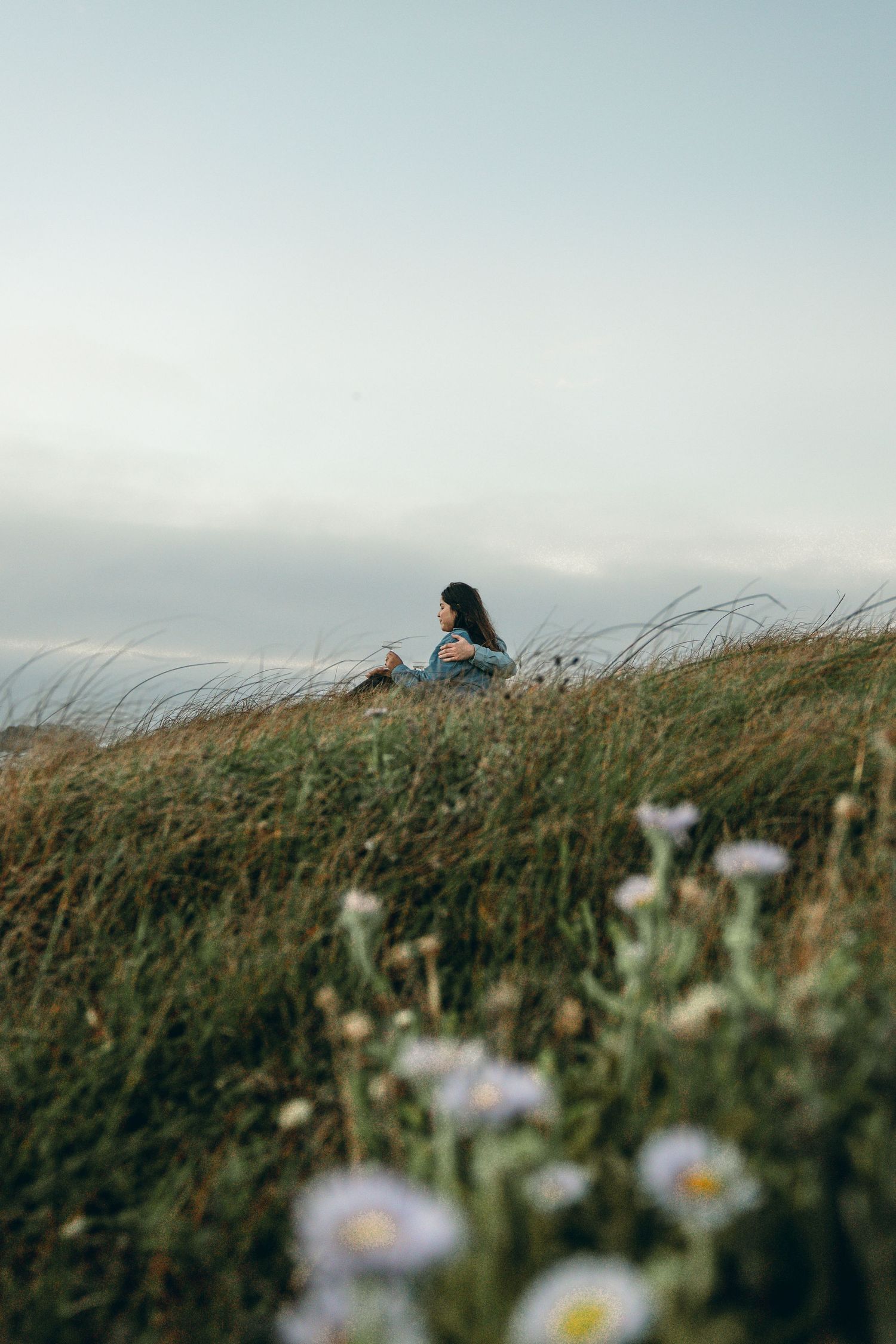 Person sitting alone on grassy hillside with wildflowers in foreground during golden hour at Asilomar Beach in Pacific Grove.
