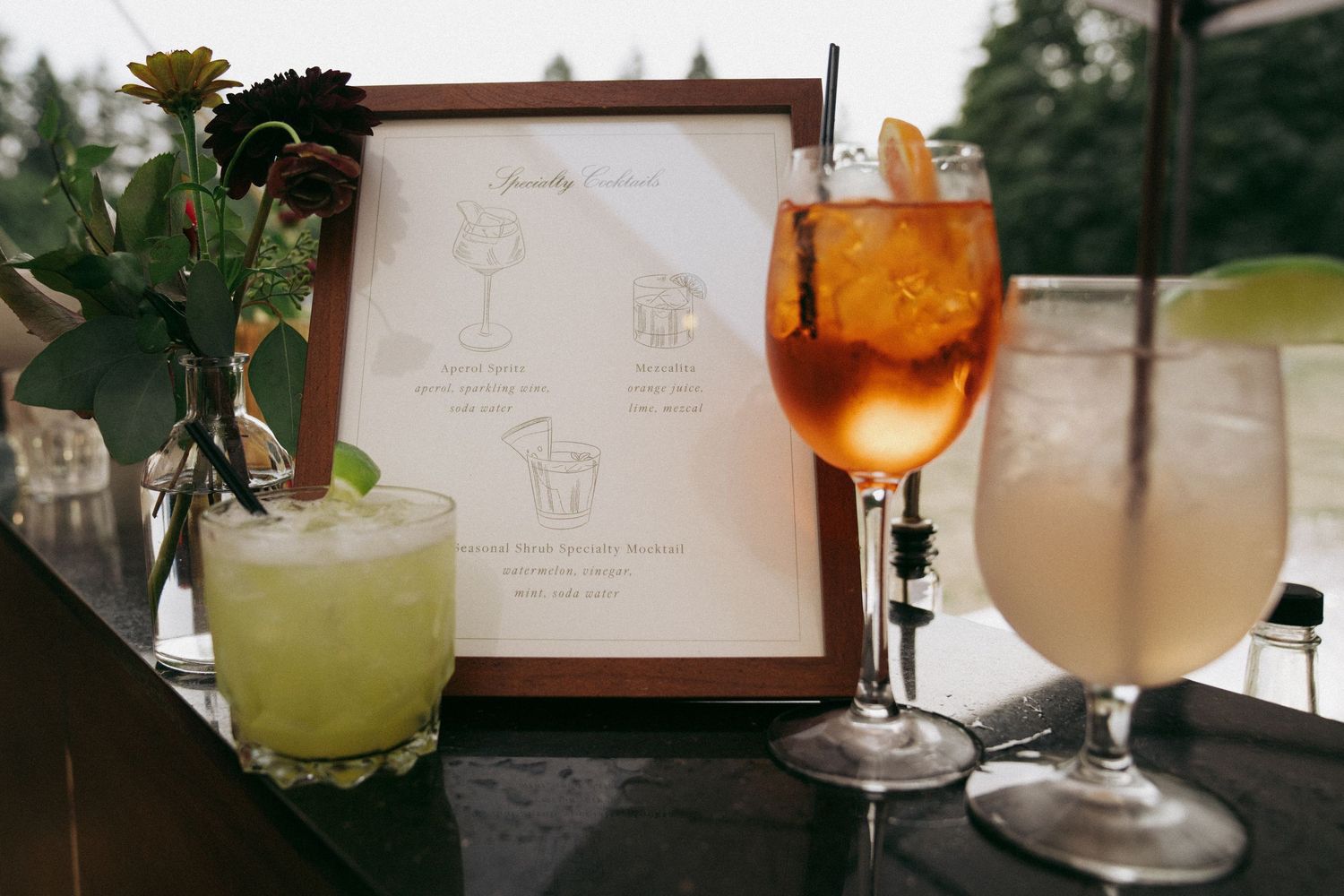 Three colorful cocktails sit on a table next to a framed menu or illustration in a restaurant setting.