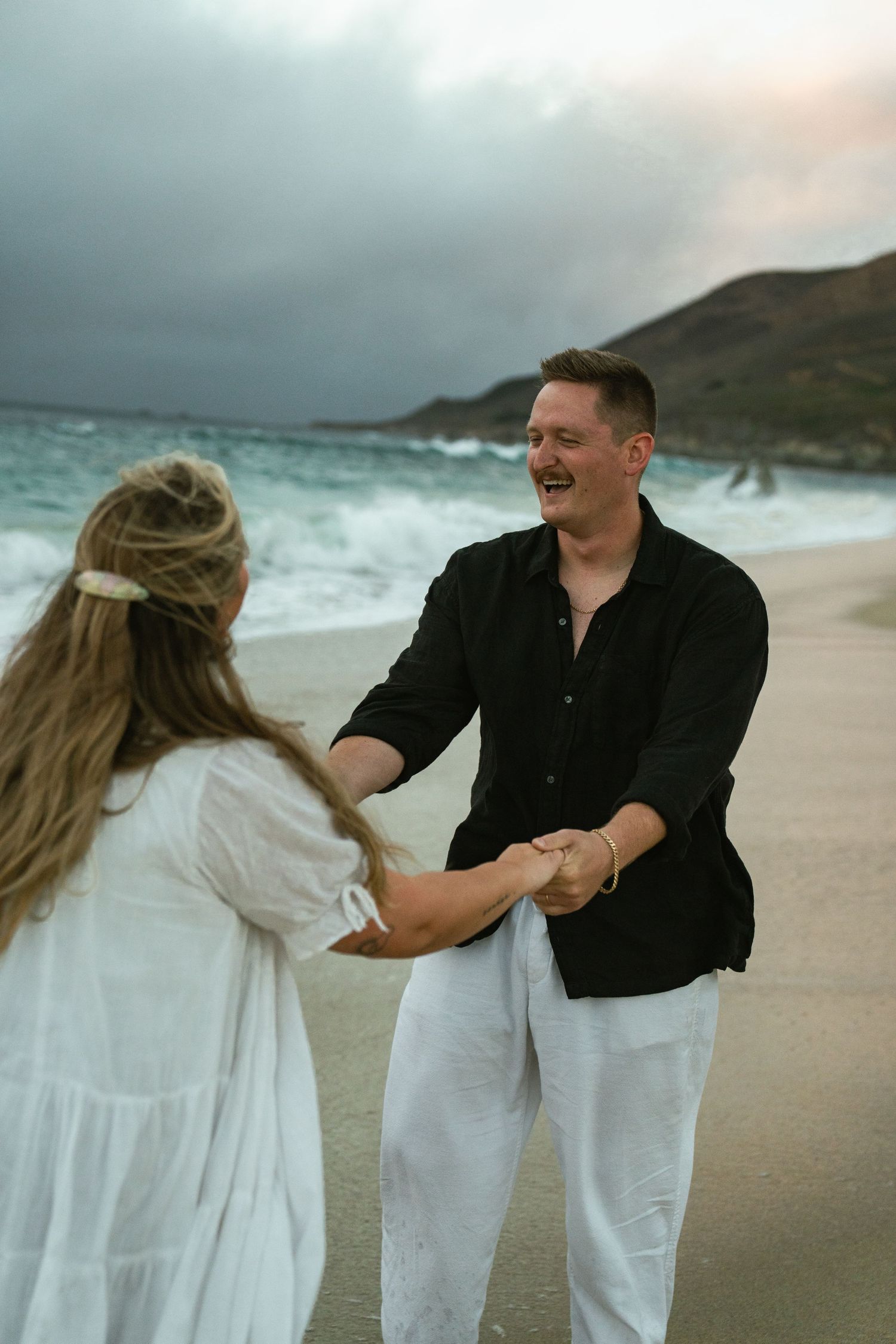A romantic beach moment unfolds as a couple embraces on a cloudy shoreline with dramatic cliffs in the background.
