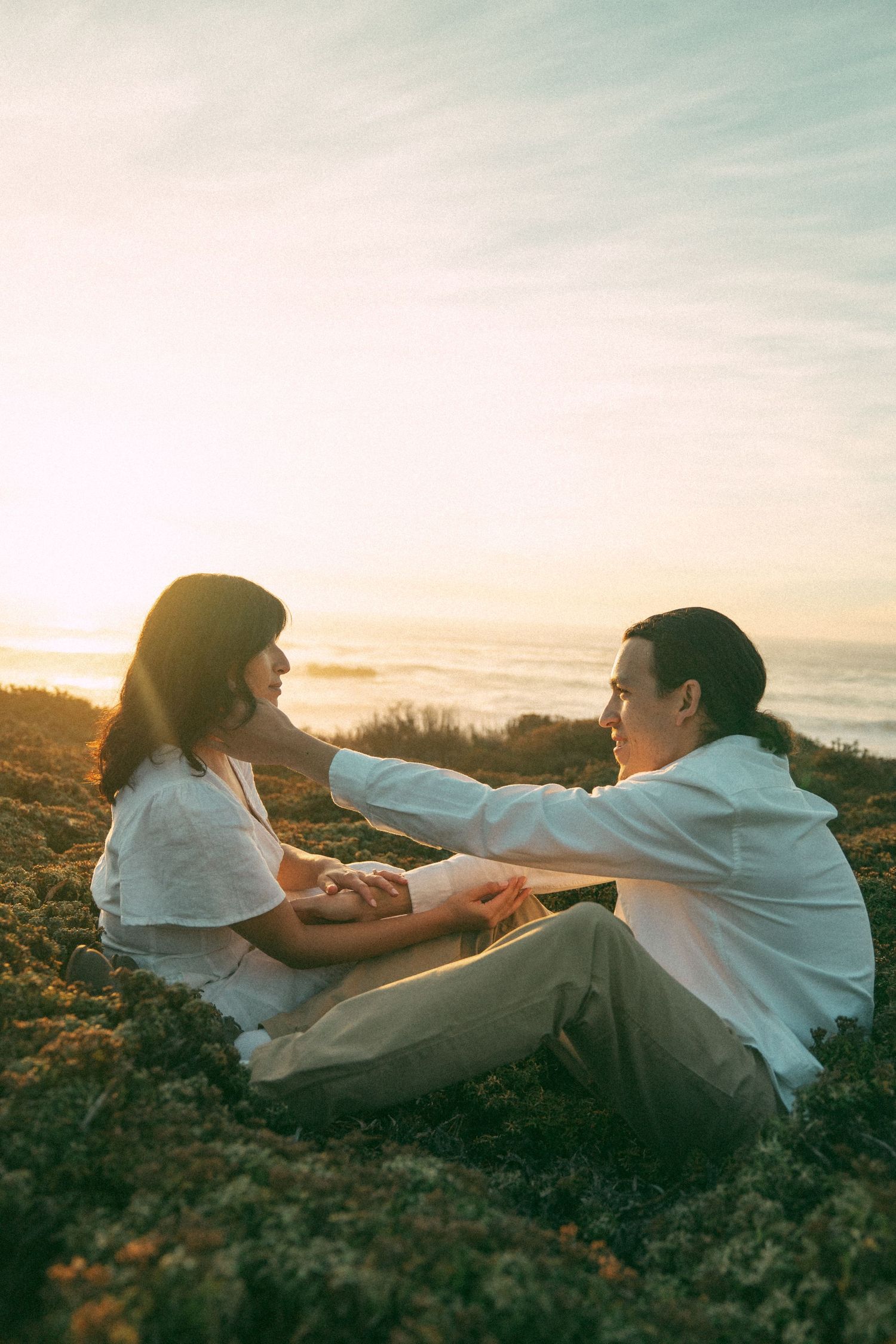 Two people in white clothing playfully interacting on a cliff at sunset by the ocean.