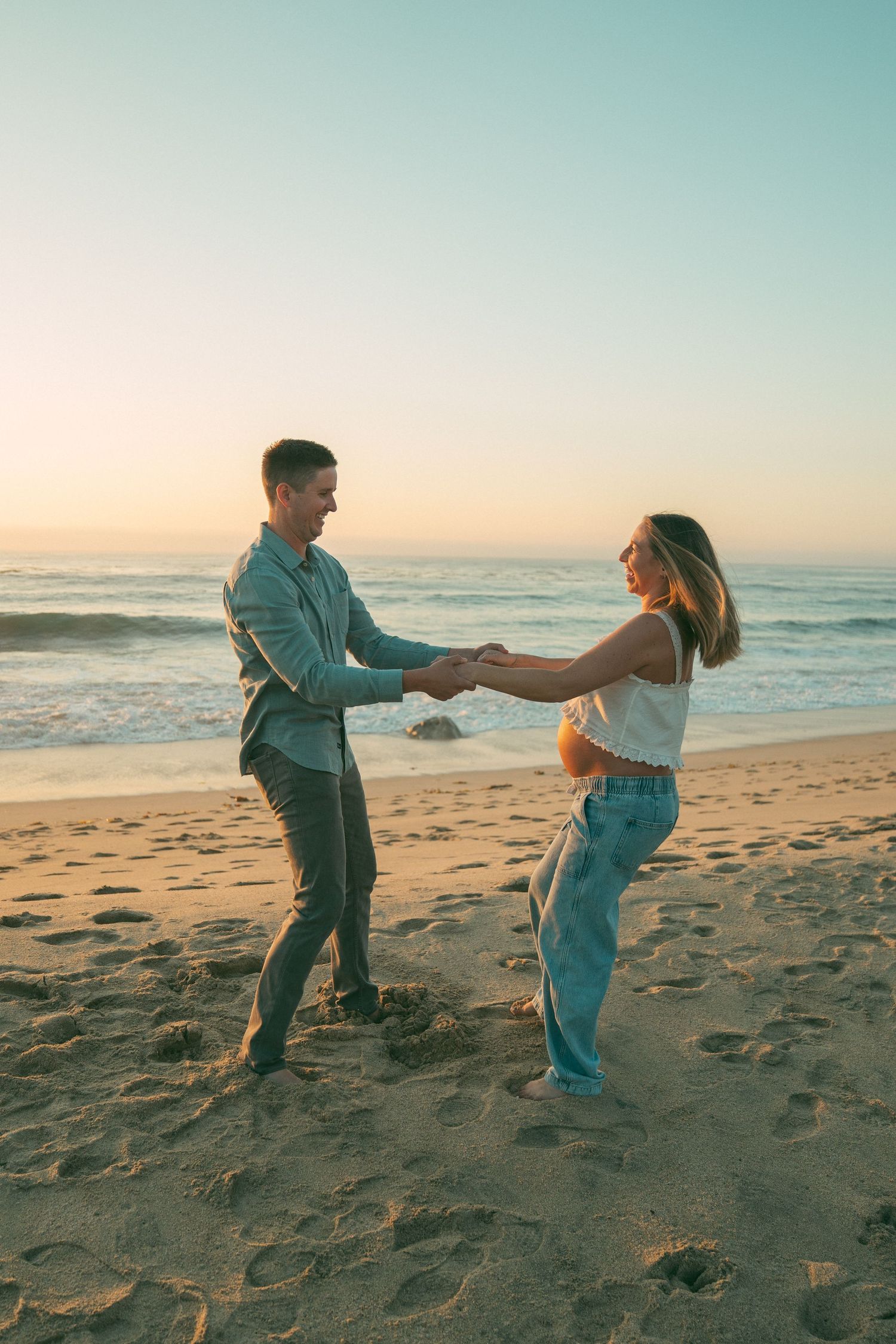 A couple dances playfully together on a sandy beach at sunset with ocean waves in the background.