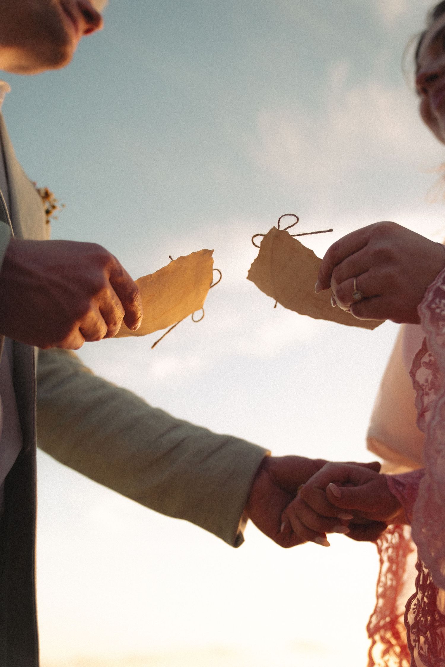 Couple exchanging handwritten vows on paper scrolls tied with twine at a romantic outdoor sunset wedding ceremony.