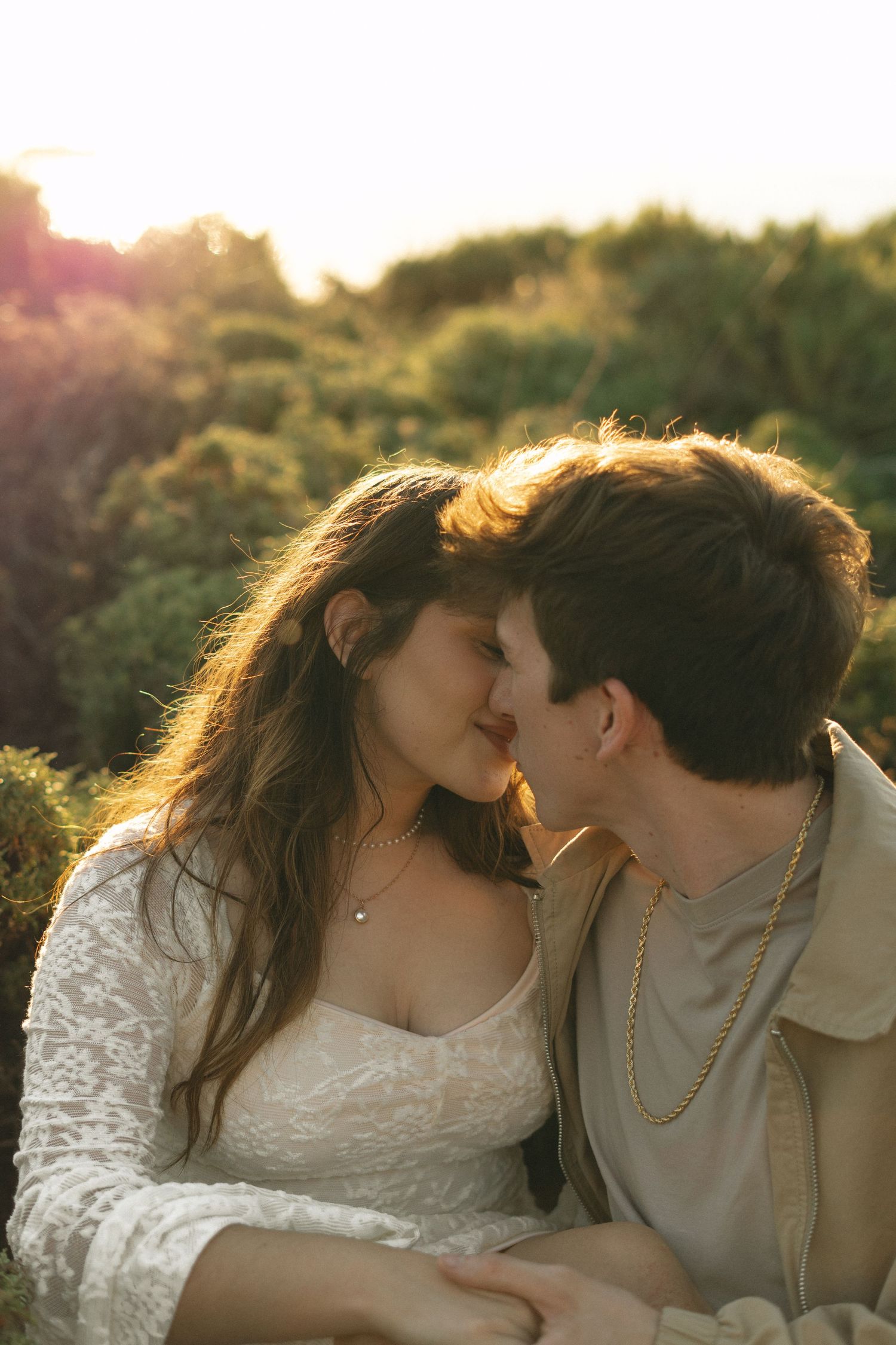 Couple sharing a romantic almost-kiss at golden hour, surrounded by lush greenery in warm sunlight.