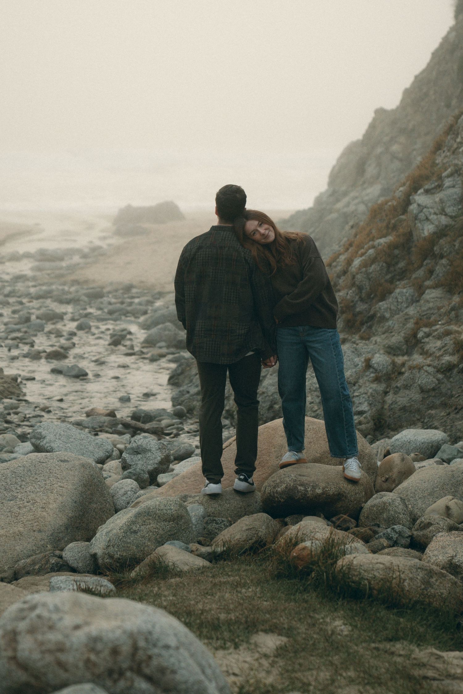 Couple walking together on rocky beach coastline during misty overcast day.