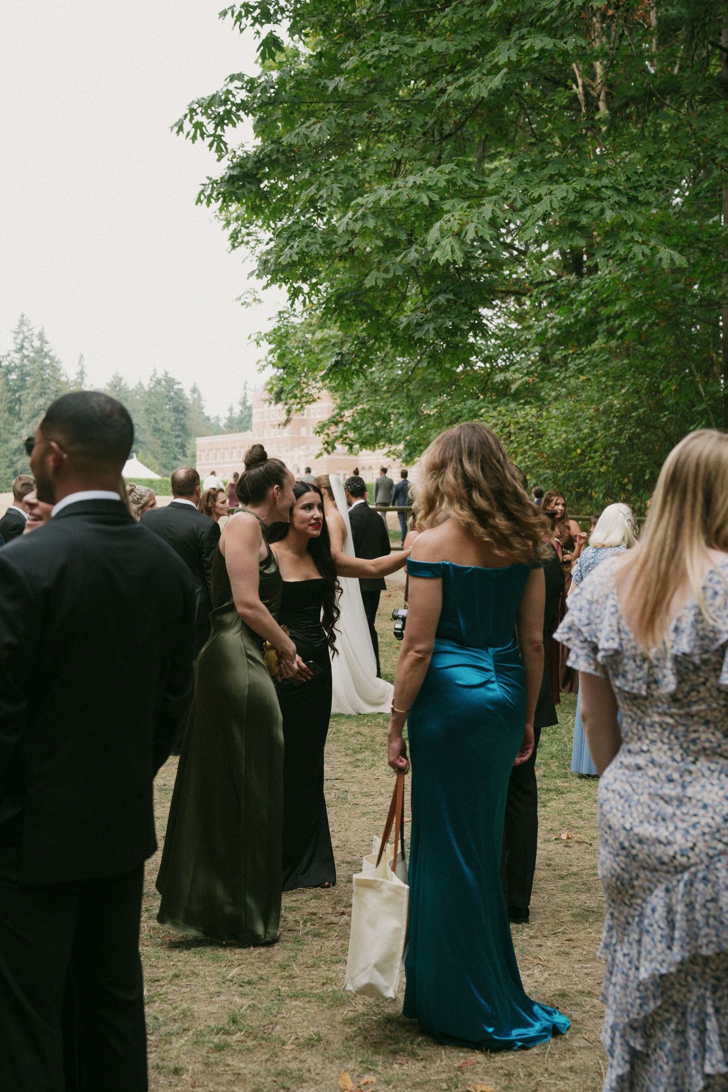 Wedding guests in formal attire gather outdoors among trees at an elegant reception.