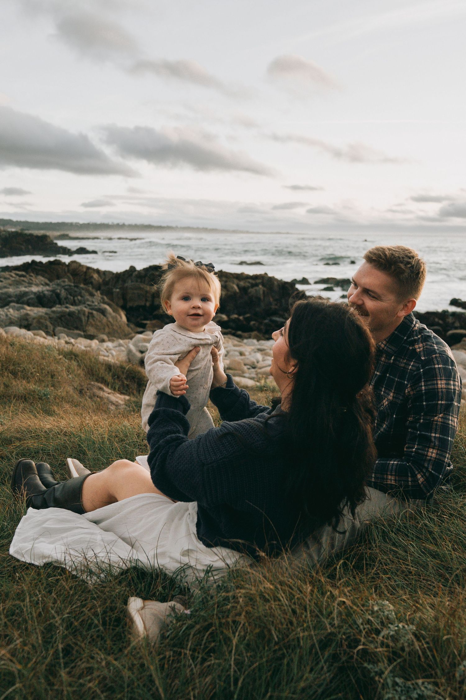 Family sharing tender moments while lounging on grass near rocky coastline at sunset.
