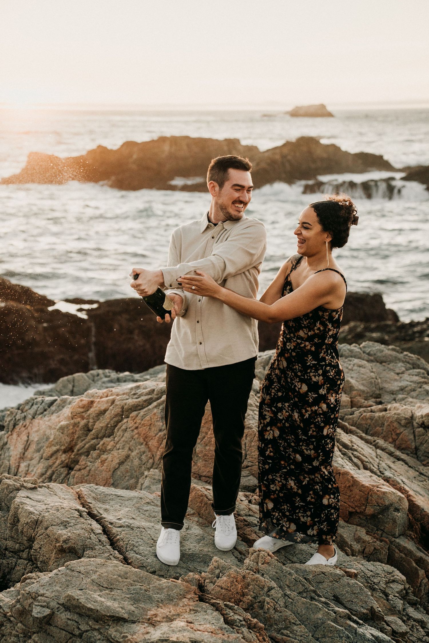 A joyful couple dances together on rocky coastal cliffs at sunset with ocean waves crashing behind them.