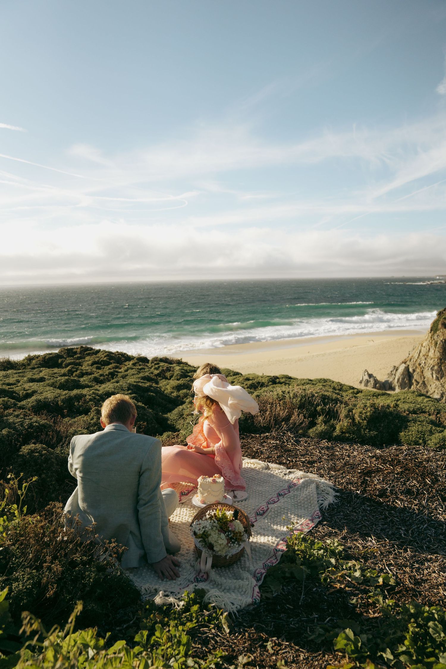 Couple sitting on a blanket overlooking a scenic beach with turquoise waves, woman in pink dress and wide hat.