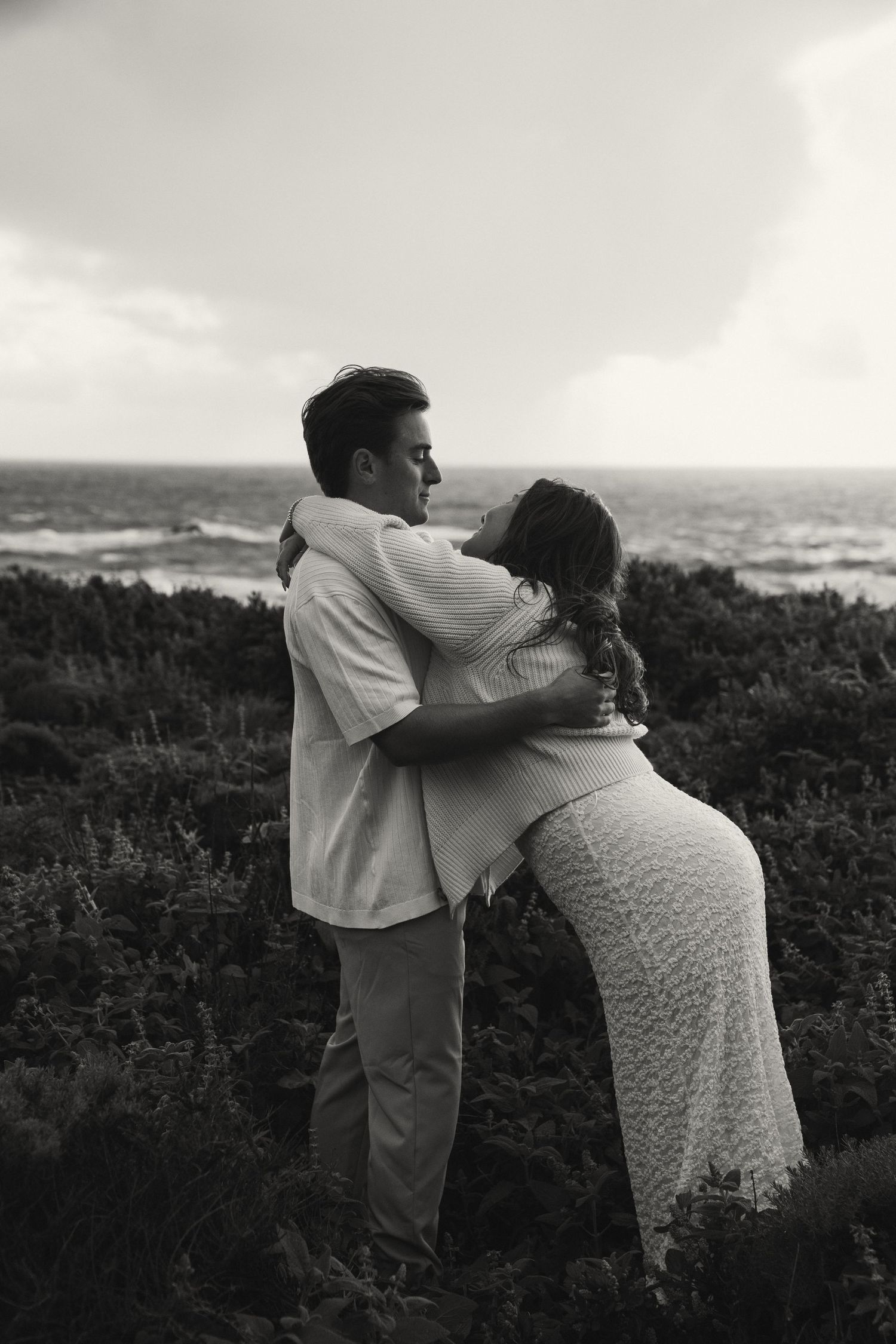 Black and white photo of a couple embracing romantically by the ocean with lush greenery and dramatic cloudy skies.
