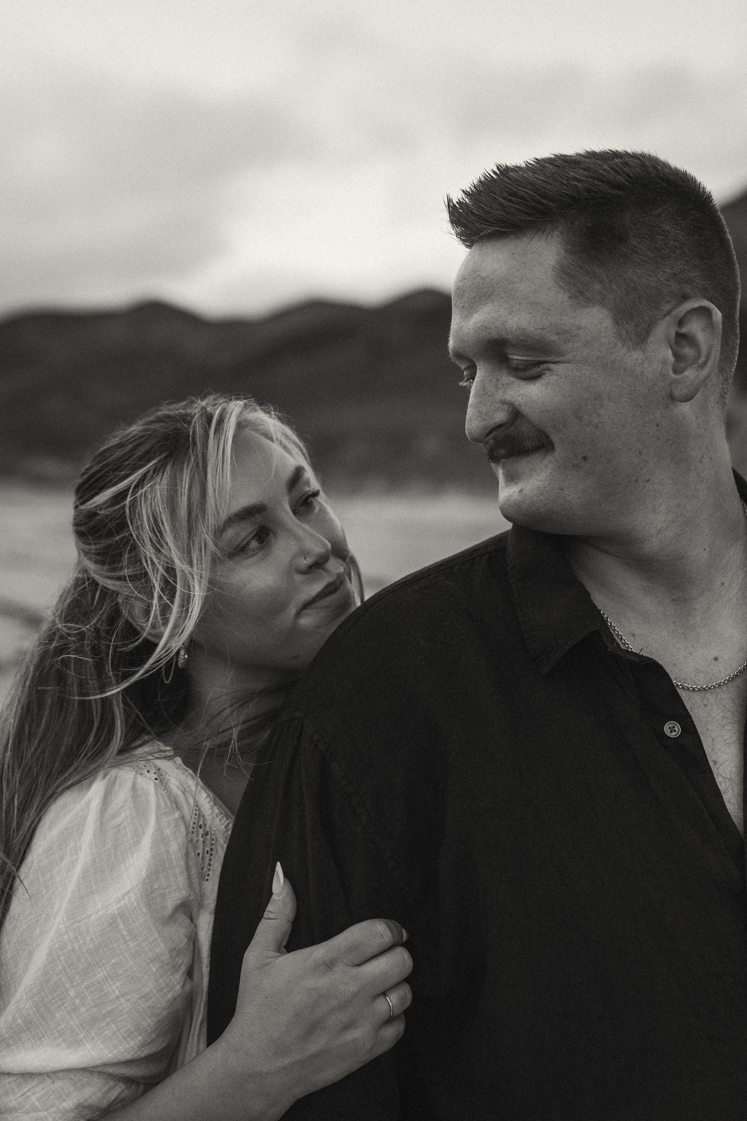 An intimate black and white portrait captures a tender moment between a couple against a mountainous backdrop.