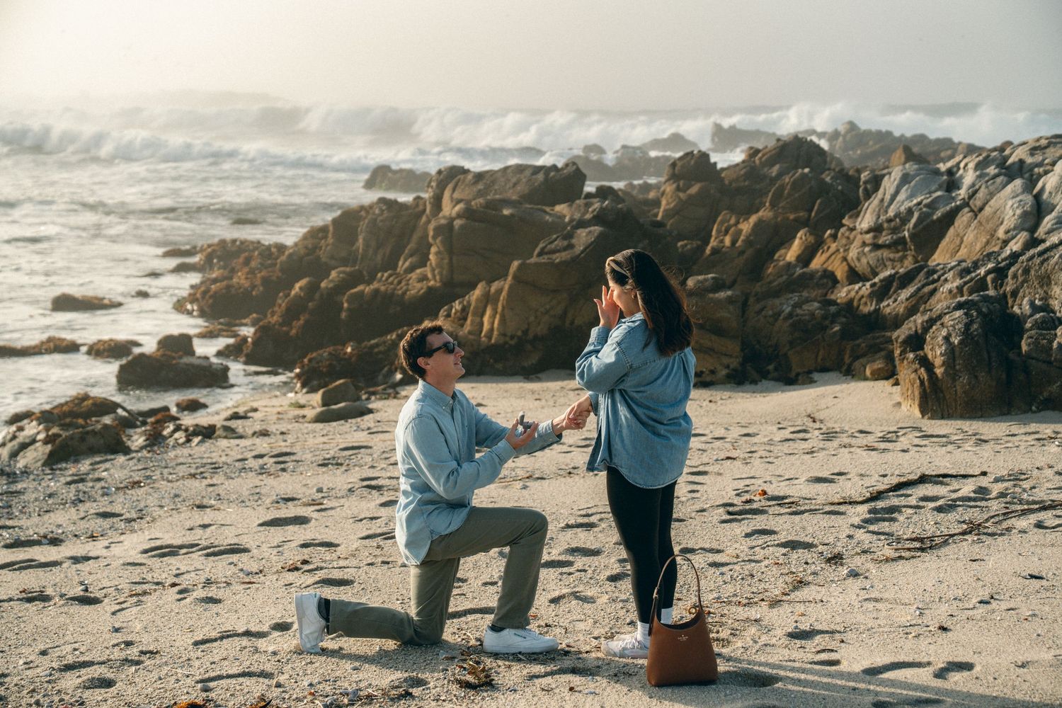 A romantic proposal moment on a rocky beach at sunset with crashing waves and coastal rocks in the background.