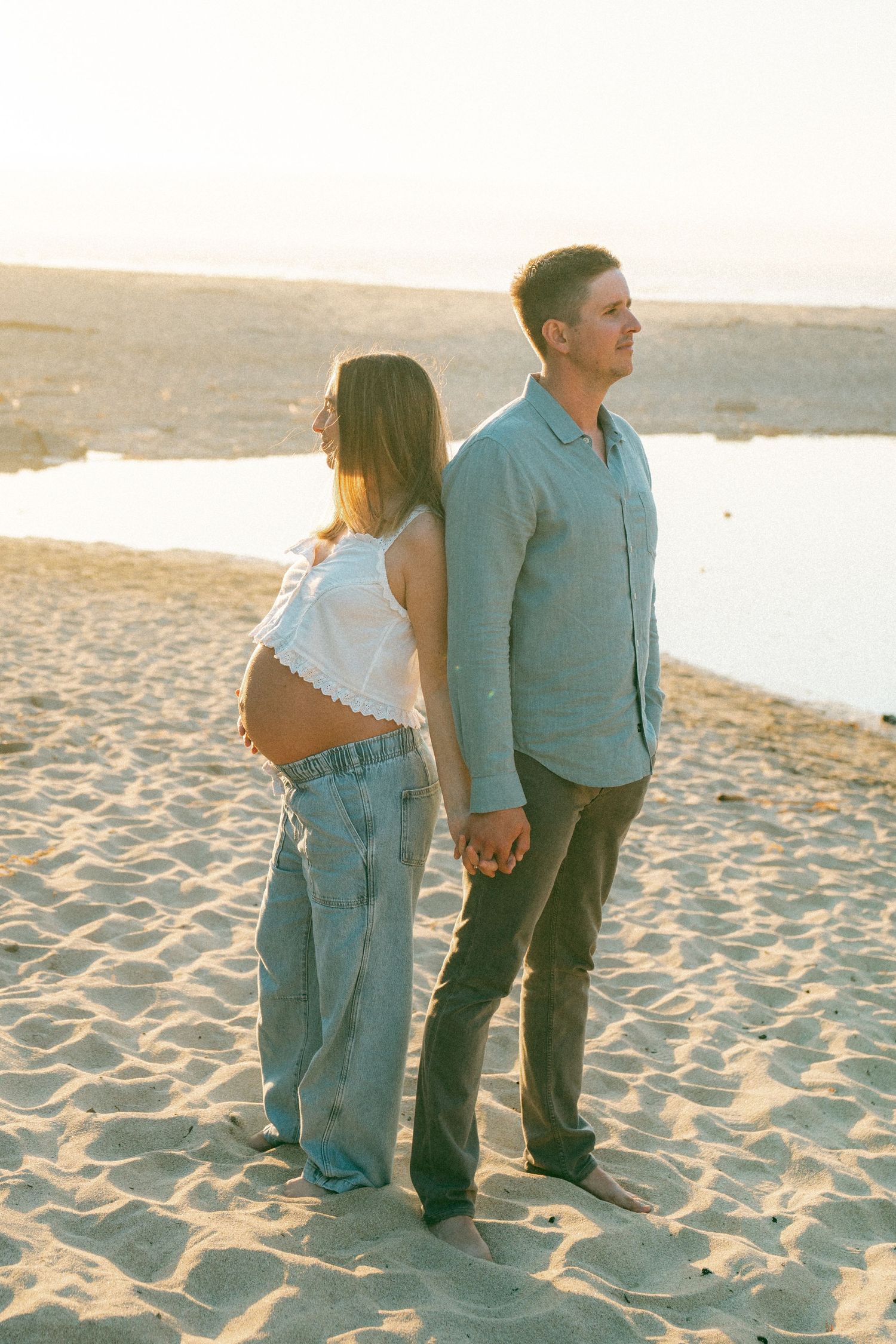 A couple holds hands while standing together on a sandy beach during golden hour.