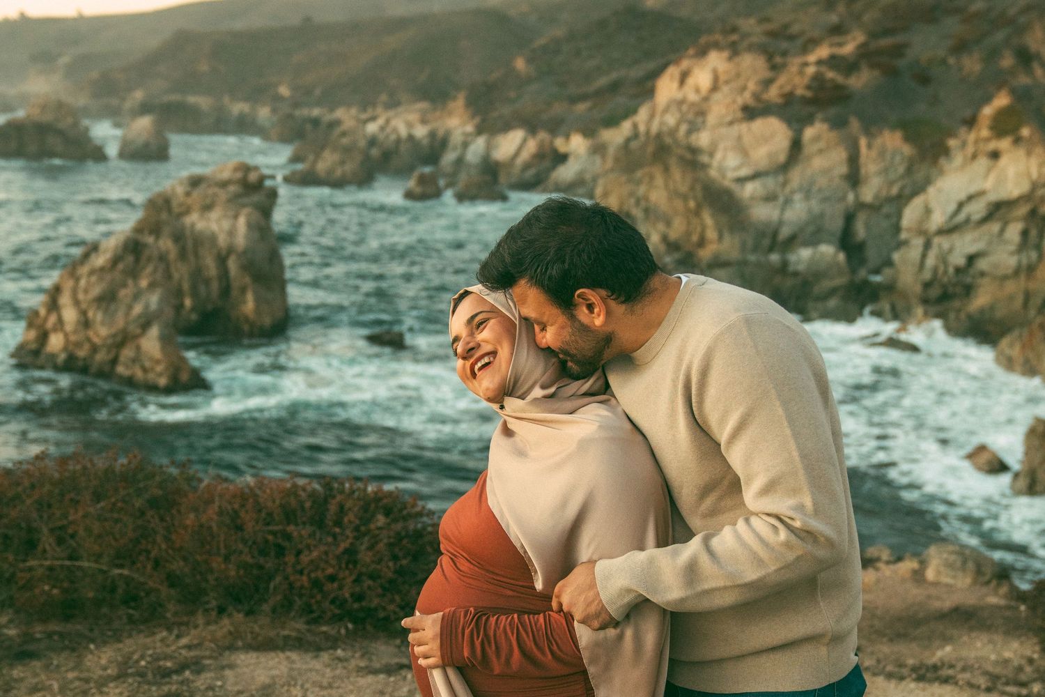 Couple embraces on rocky coastal cliffs overlooking dramatic ocean waves at sunset.