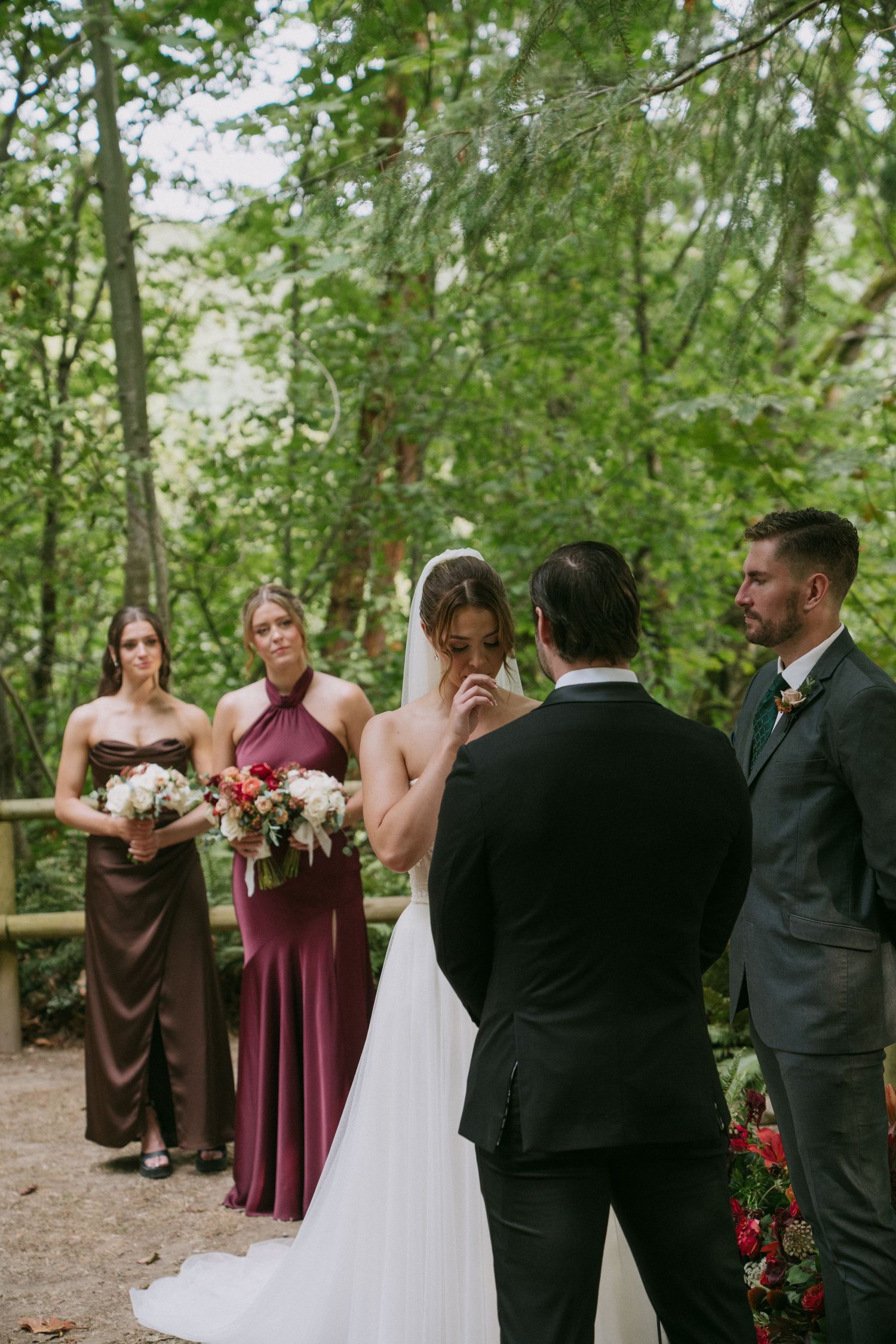An outdoor wedding ceremony in progress with bridesmaids in burgundy and brown dresses among trees.