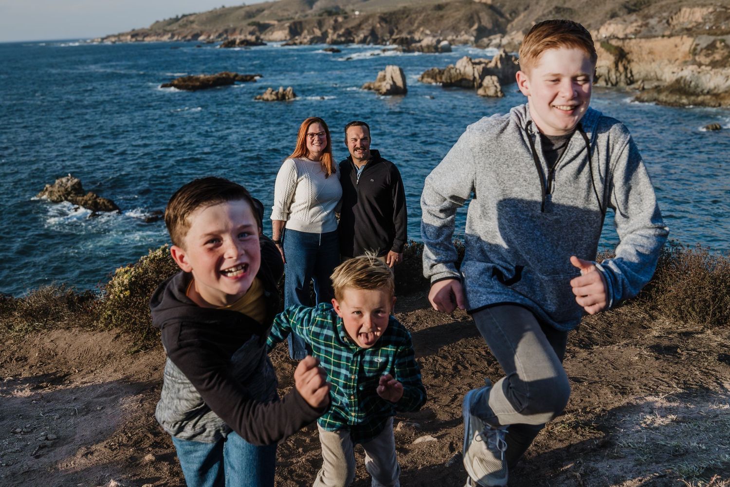 A group capers playfully along a scenic coastal path with rugged cliffs and blue ocean waves in the background.