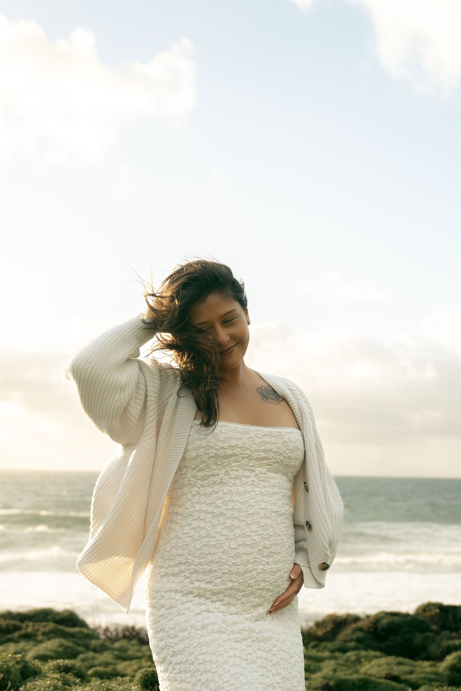 Pregnant woman in white dress and cardigan smiling at beach, ocean waves and cloudy sky in background.