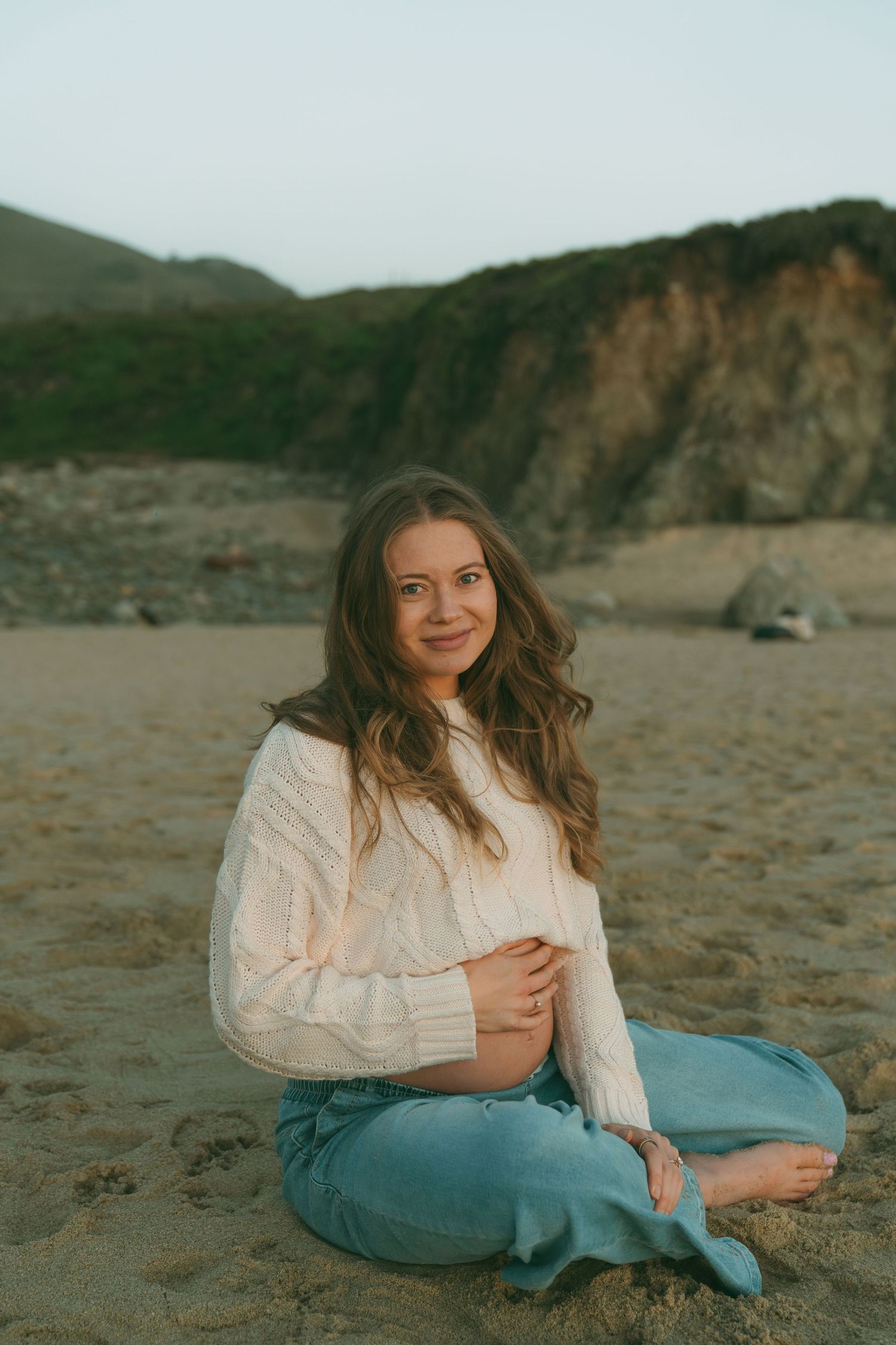 Pregnant woman sitting cross-legged on sandy beach, wearing white sweater and jeans, smiling with rocky cliffs behind her.
