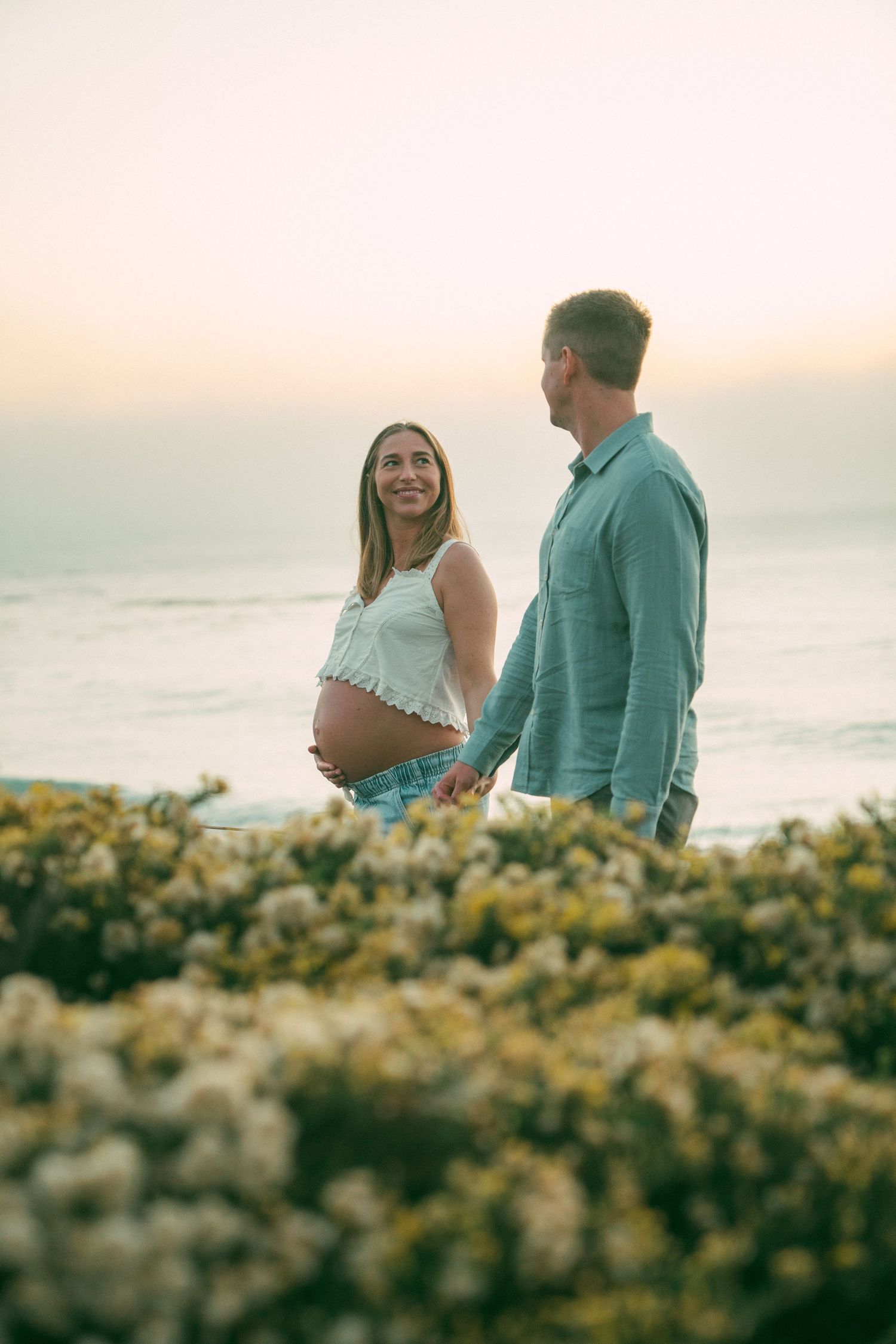Couple sharing intimate moment among coastal flowers during sunset photoshoot.