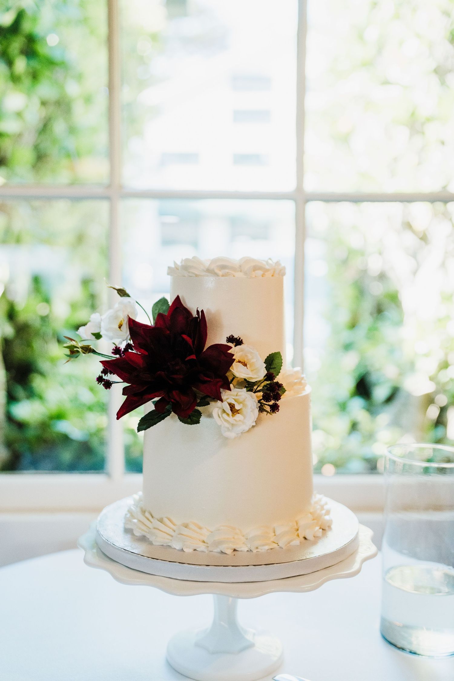 Elegant white two-tier wedding cake decorated with burgundy and white flowers on a pedestal stand by a bright window.